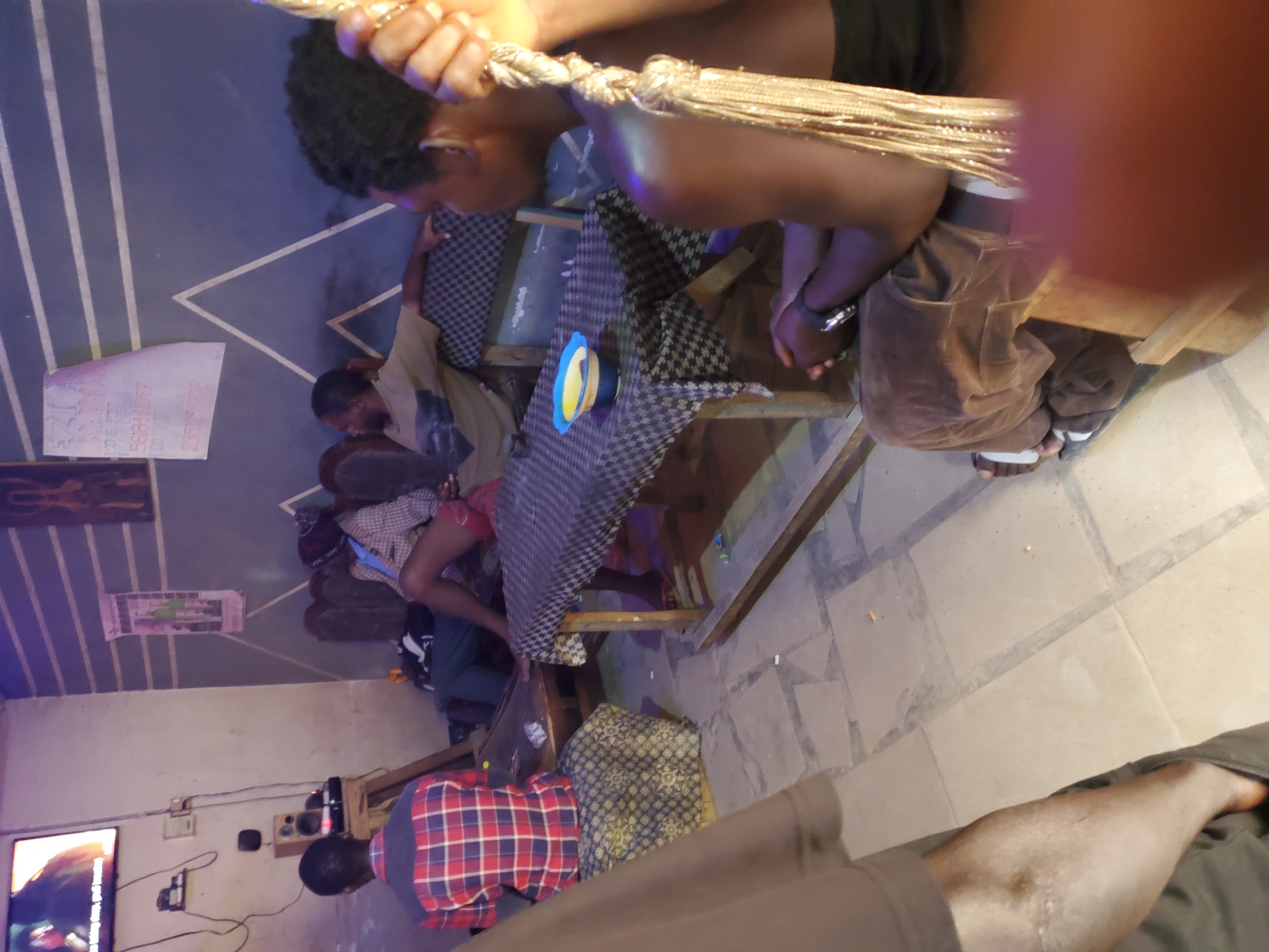 The image captures an indoor scene within a dimly lit establishment, possibly a bar or lounge, in Jos, Nigeria. Four individuals are present, seemingly engaged in casual interaction. The ambiance suggests a relaxed, perhaps informal gathering.
On the left, a person wearing a red and blue checkered shirt sits facing away from the camera, their back to a television screen mounted on the wall. Next to them, another individual in a patterned outfit is seated, their legs crossed. Further to the right, two other individuals are visible. One, in a light-colored shirt, leans back, seemingly in conversation or watching something. The fourth individual, in the foreground and facing towards the right, has their back to the camera and is holding onto decorative beaded strands.
A low table is positioned in the center of the frame, covered with a checkered cloth. On the table sits a blue and yellow bowl. The floor is tiled with a geometric pattern. The walls are decorated with horizontal stripes and geometric shapes in shades of blue. The overall setting appears to be a communal space, with elements suggesting a place for leisure and socializing.