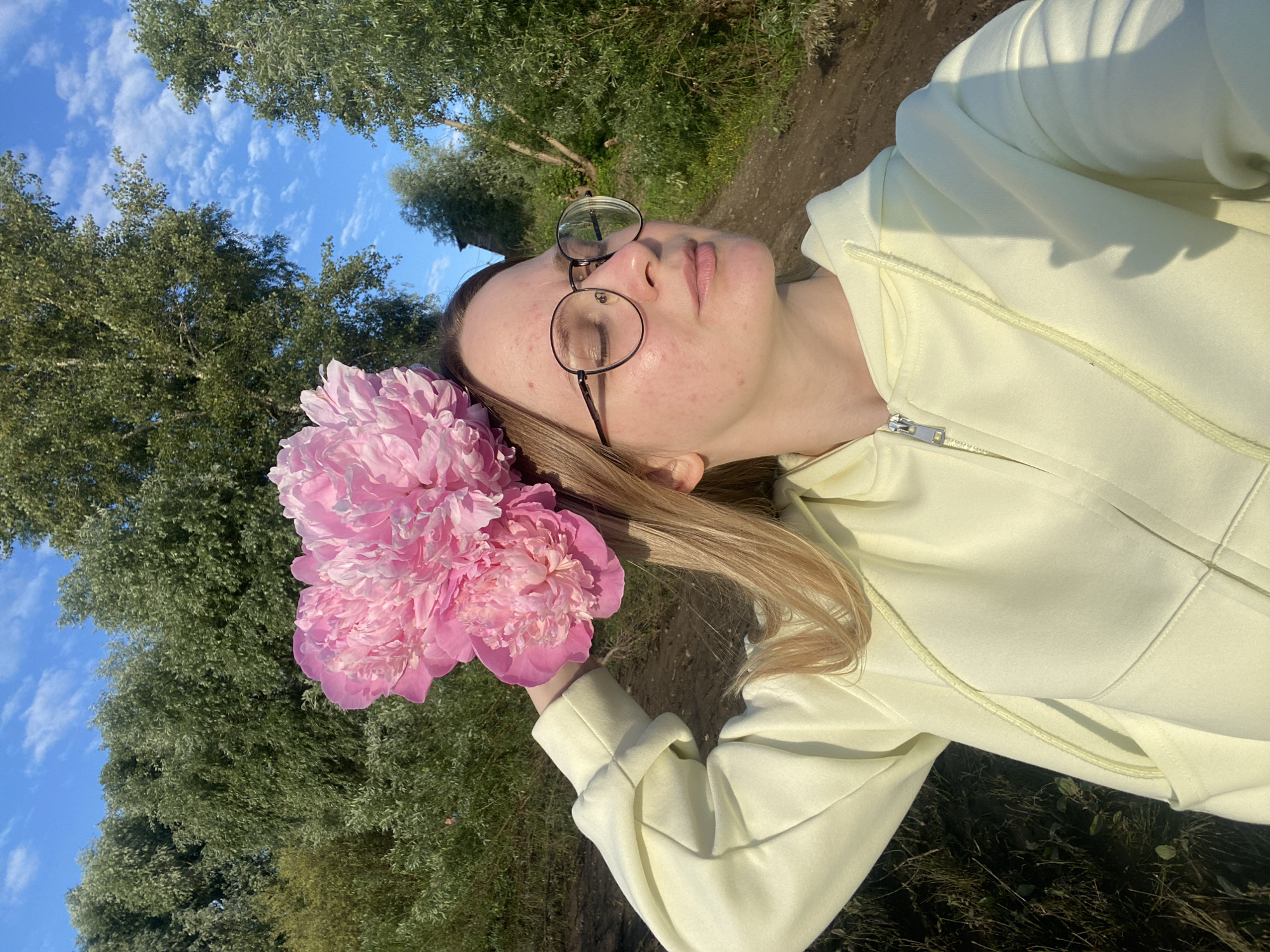 This media file shows a young woman lying down outdoors, with her head tilted back and eyes closed. She is wearing round, wire-rimmed glasses and a light yellow hoodie. In her right hand, she holds a bouquet of vibrant pink peonies. Her blonde hair is spread out around her head.
The background consists of lush green trees, with a bright blue sky dotted with white clouds visible above. The ground appears to be a mixture of soil and dry grass, suggesting a natural or semi-rural setting. The lighting indicates it is daytime, likely late afternoon or early evening given the warm sunlight casting shadows.
The woman's posture and closed eyes convey a sense of relaxation and peace. The overall atmosphere is serene and natural, enhanced by the beauty of the peonies and the surrounding greenery. There are no visible text elements in the image. The context provided places this scene in Chaikovsky, Russia.