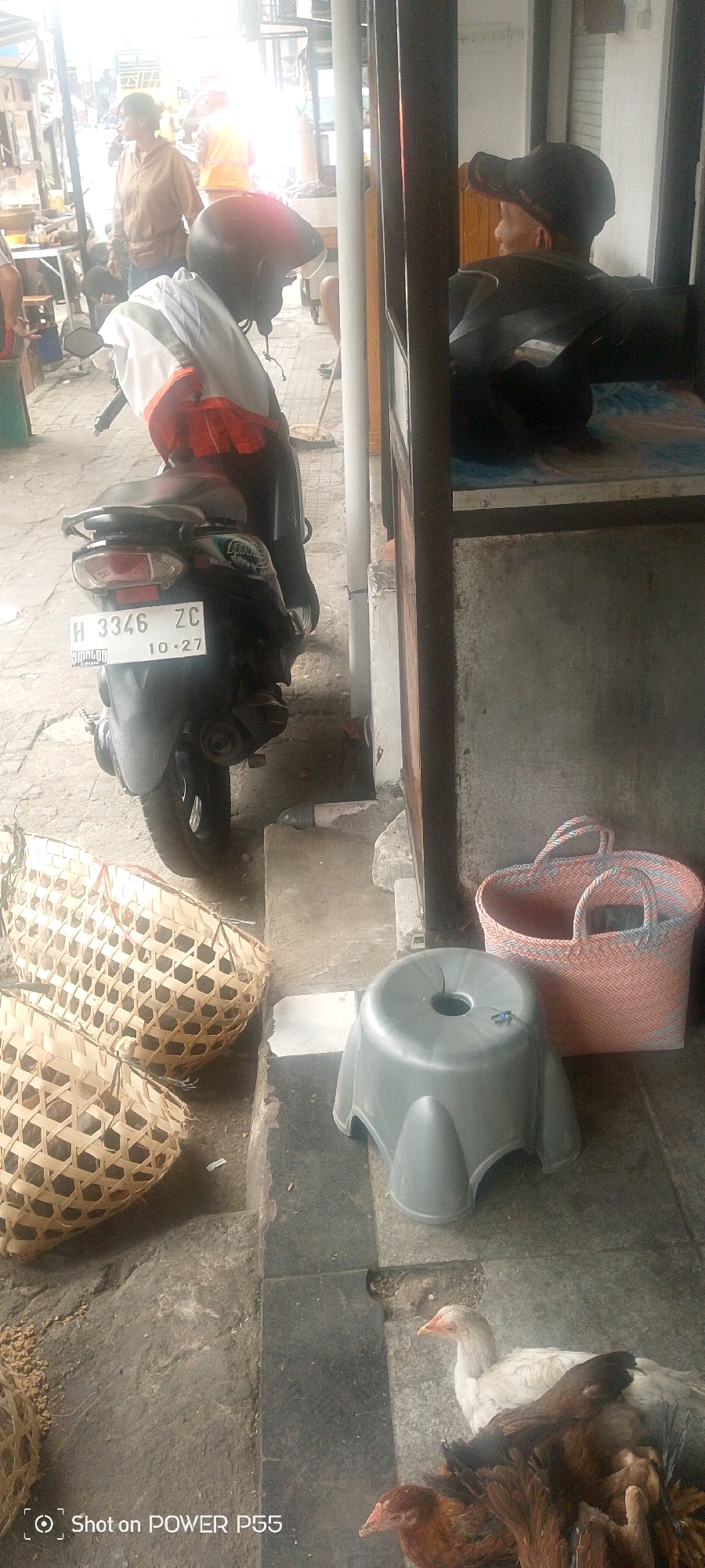 A motorcycle with a license plate that reads "H 3346 ZC 10-27" is parked on a sidewalk in front of a market stall. A person wearing a helmet and an orange vest is seated on the motorcycle. Two woven baskets are placed on the ground next to the motorcycle. A gray plastic stool is situated in front of a pink and blue woven bag. Two chickens are visible on the ground near the stool and bag. In the background, a woman in a brown jacket and a person wearing a yellow vest are seen near food stalls. Another person wearing a black cap and jacket is sitting. The scene appears to be an outdoor market in a city.