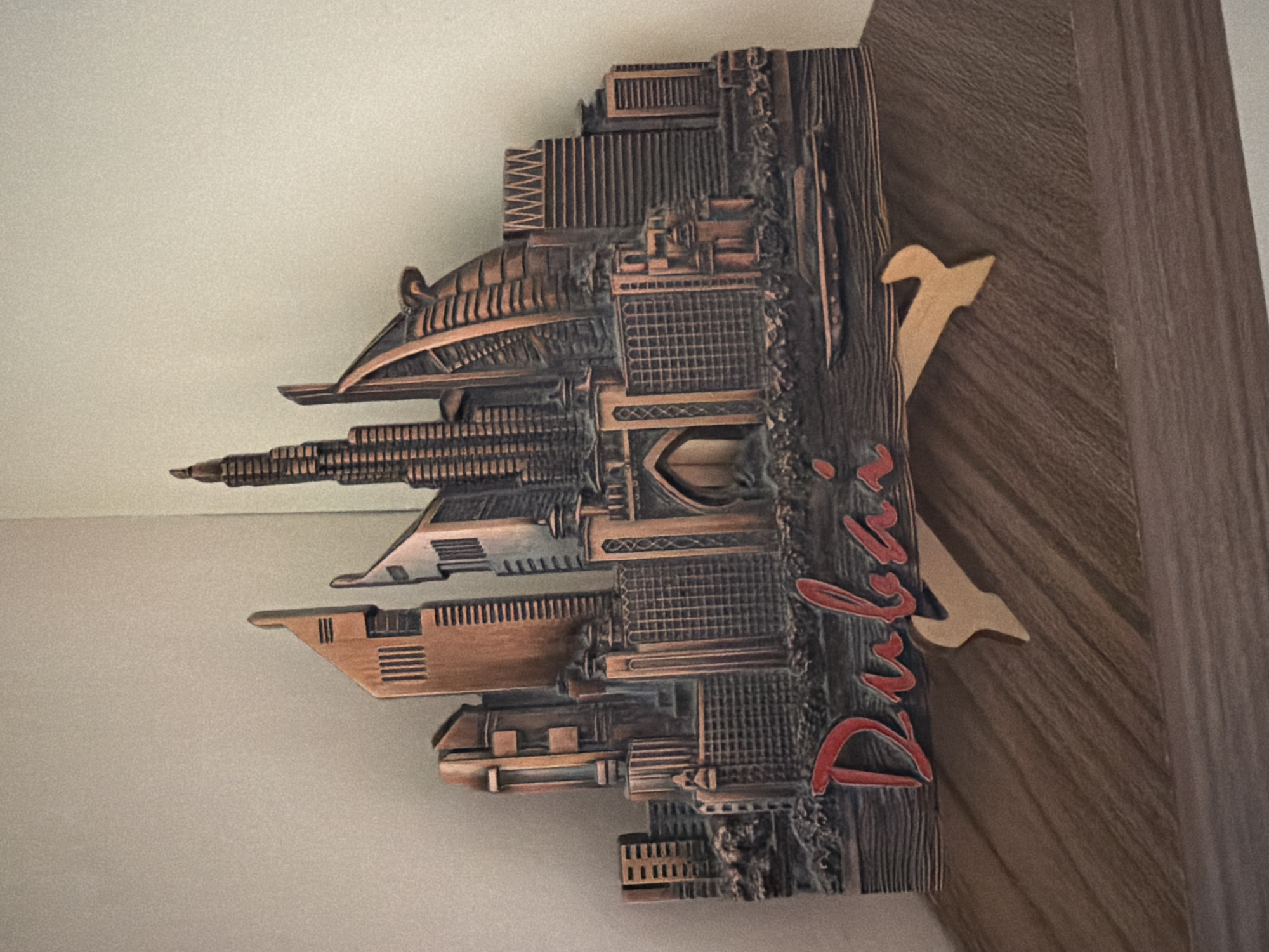 A copper-colored 3D replica of the Dubai skyline, featuring several iconic skyscrapers and buildings, is displayed on a wooden surface. The replica includes the Burj Khalifa, Burj Al Arab, and other modern architectural structures. The word "Dubai" is written in red, stylized script below the skyline. The replica is supported by two light brown, Y-shaped stands. The background is a plain, light-colored wall, possibly painted. The scene is lit from the front, casting subtle shadows. There are no people present in the image. The replica is positioned on a shelf or table with a wood-grain texture. The location is not Pazardzhik, Bulgaria.