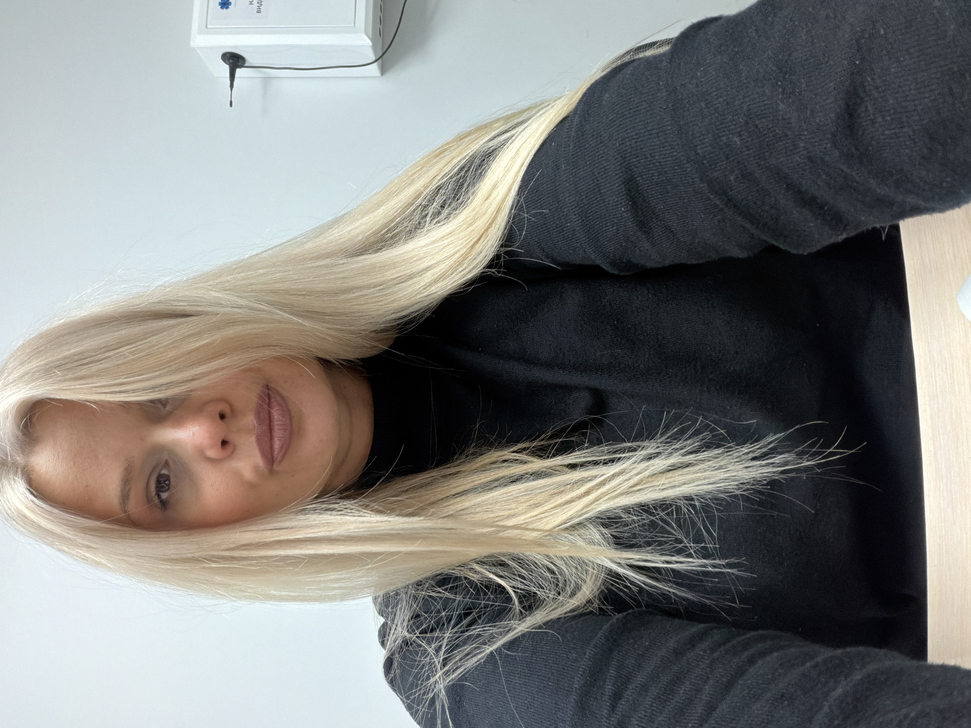 A woman with long blonde hair is visible in the image. She is wearing a black turtleneck sweater. The background consists of a white wall with a white device mounted on it. The image appears to be taken indoors. There is no information about Sarapul, Russia.