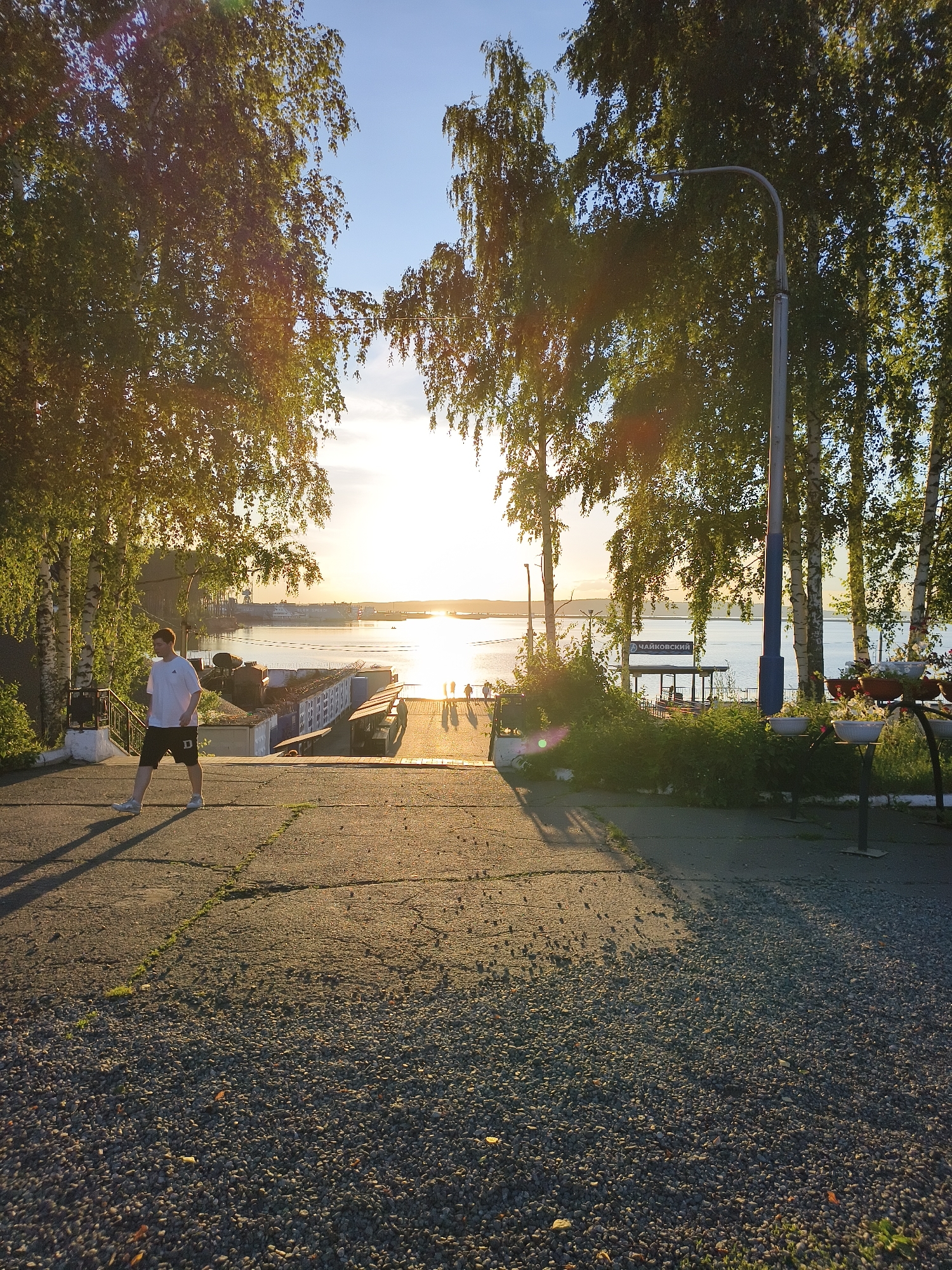 A young man wearing a white t-shirt and black shorts walks down a sloped asphalt path with gravel on the sides. He is casting a long shadow. Behind him, trees with green leaves frame a view of a calm body of water reflecting the golden sunlight of a sunset. In the distance, buildings and boats are visible on the water. A street lamp stands to the right of the frame. The scene appears to be a waterfront area in a city.
