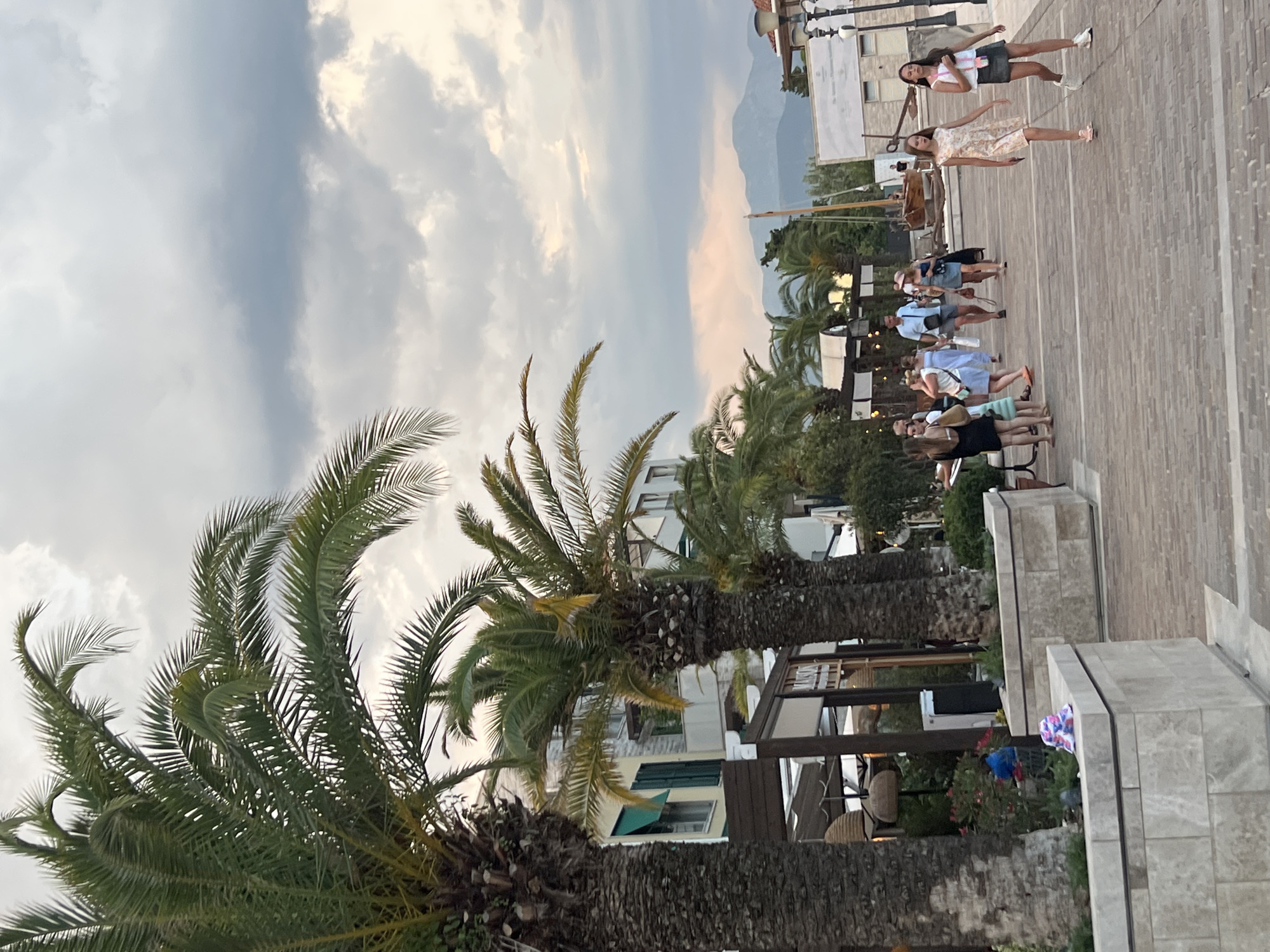 A group of people are walking along a paved walkway lined with palm trees. The scene appears to be a waterfront promenade in a Mediterranean town, with buildings and boats visible in the background. The sky is cloudy, suggesting it might be late afternoon or early evening. The people are dressed in casual summer clothing, and some are carrying bags. The overall mood of the image is relaxed and leisurely. There are no visible signs or text in the image.