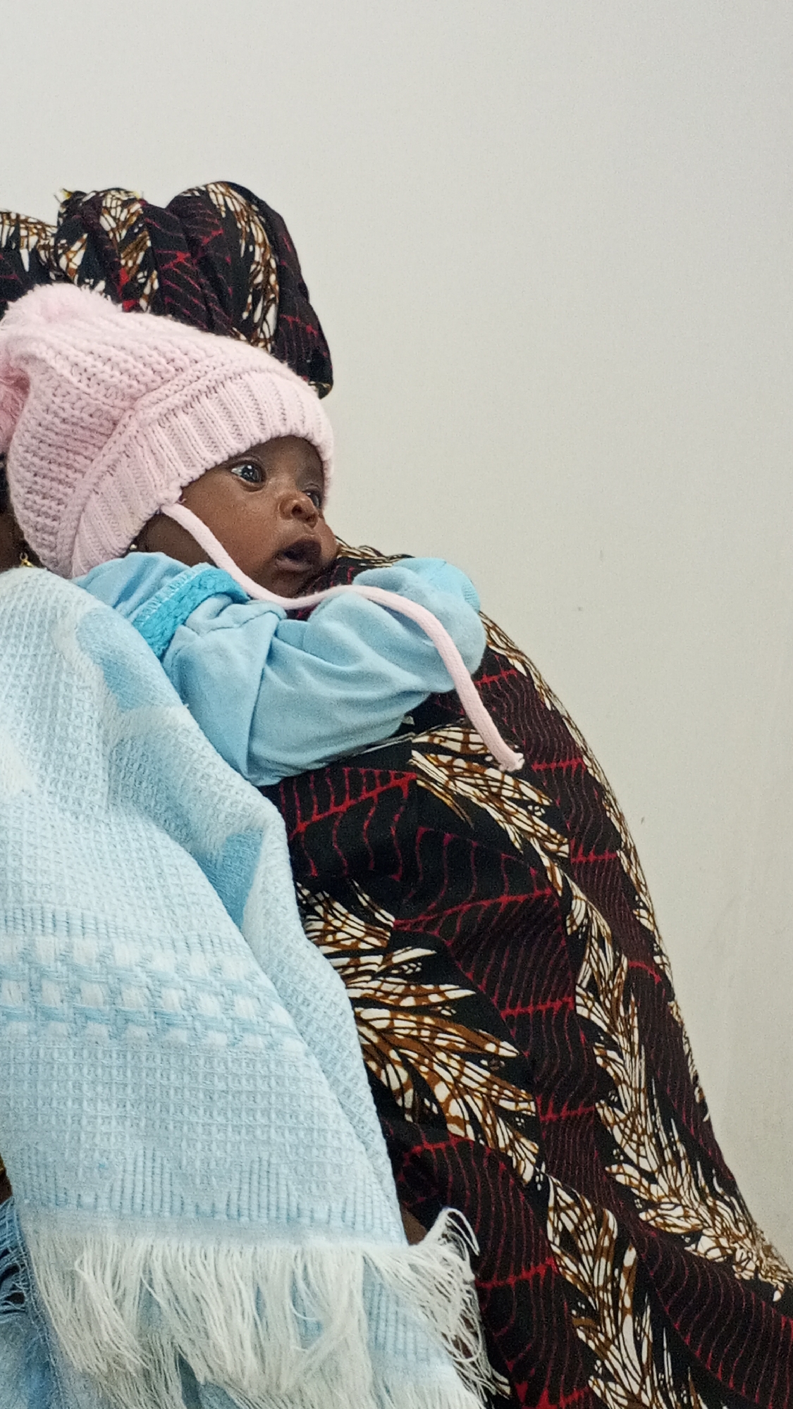 A baby wearing a pink knitted hat and a light blue shirt is being held in someone's arms. The person holding the baby is wearing a patterned fabric. A blue blanket with white fringe is also visible. The background is a plain, light-colored wall. There are no readily apparent contextual details to establish that this image was taken in Lokoja, Nigeria.