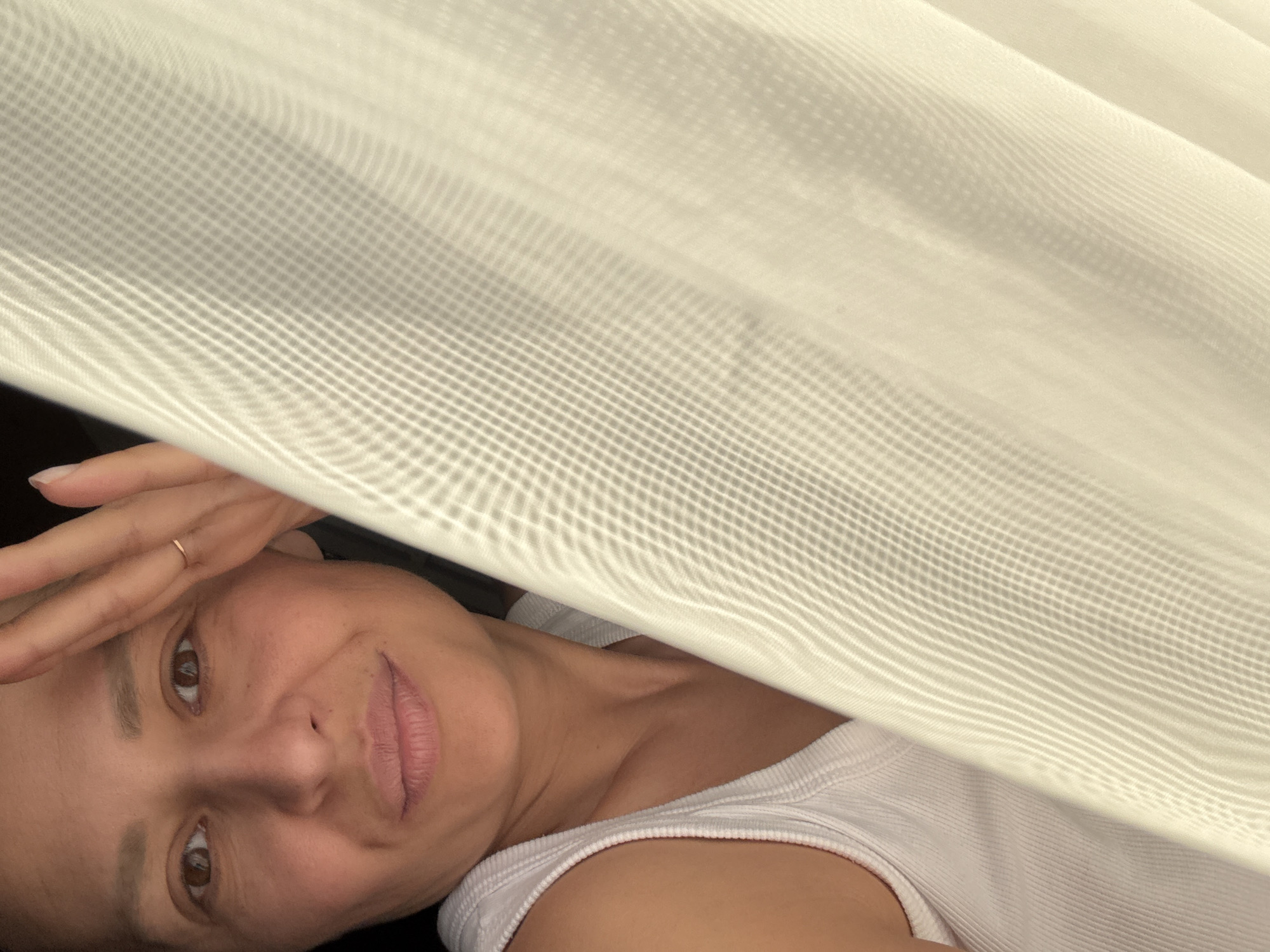 A woman with brown eyes and light brown hair is positioned behind a semi-transparent curtain, possibly sheer fabric, with a grid-like pattern. She is wearing a white tank top and a gold ring on her left ring finger. Her right hand is raised to her forehead. The location is not identifiable as Chaikovsky, Russia from the visual information provided.