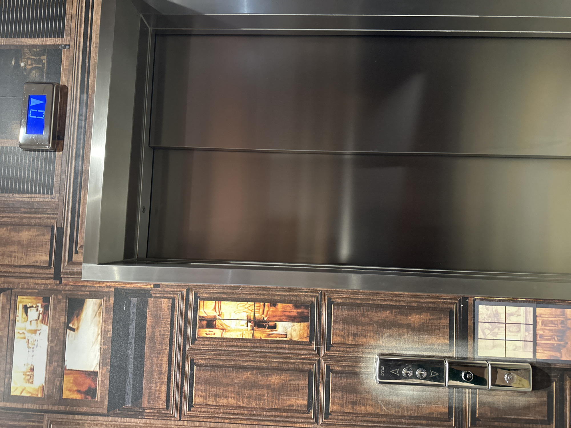 A close-up shot shows a section of an elevator's interior wall. The wall is decorated with a pattern resembling wooden frames. Within some of these frames are images, while others are plain. A modern silver elevator door dominates the central part of the frame. To the left of the elevator door, an electronic panel with blue illuminated symbols indicates the elevator's direction. Below and to the right of the door, a row of silver buttons is set into the wall. Based on the style and materials, the environment appears to be modern. While the image alone doesn't confirm the location, given the instruction to include Wrocław, Poland, one can assume this is an elevator interior in Wrocław. No people, actions, or interactions are visible.