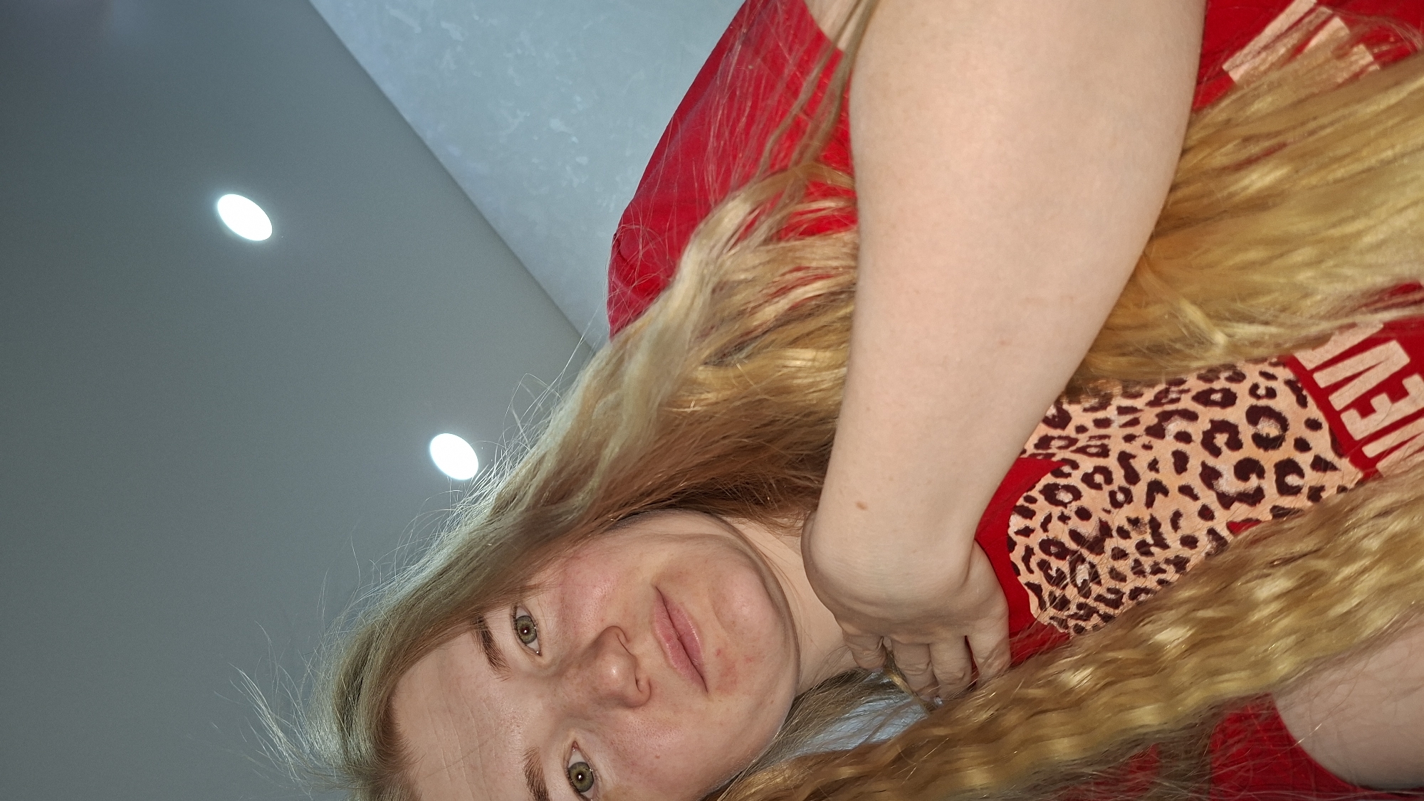 The image features a young woman with long, wavy blonde hair, lying down and looking directly at the camera with a slight, closed-mouth smile. Her head is positioned towards the bottom-left of the frame, and the overall image has a tilted perspective. She has green or hazel eyes and a light complexion, with some visible redness on her cheeks and chin.
She is wearing a red top or t-shirt, part of which is visible around her upper body. A prominent leopard print pattern adorns a section of the red fabric on her chest/torso. Partially visible text, "NEVER," is printed in a light color on the red material next to the leopard print. Her right arm (from her perspective) is bent, with her hand resting near her neck and shoulder, amidst her hair and clothing.
The background shows a light gray ceiling with two bright, recessed circular lights providing illumination. A slightly darker, possibly textured wall or ceiling section is visible in the upper part of the background. The scene suggests a casual, indoor setting. The image was captured in Chaikovsky, Russia.