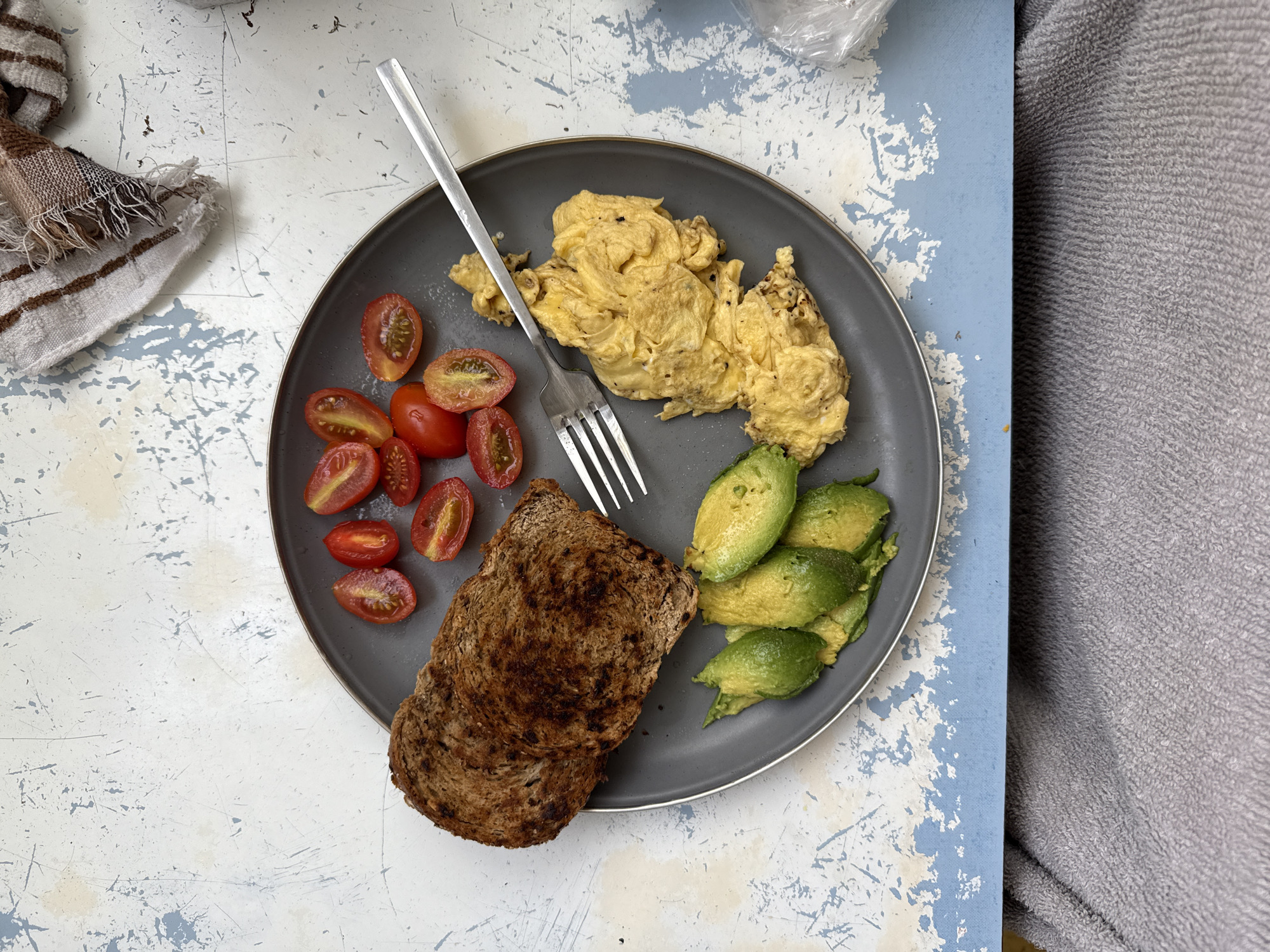A plate of food is centered on a distressed blue and white surface, with sections of the surface showing through. The plate, dark gray in color, contains scrambled eggs, sliced avocado, halved cherry tomatoes, and two slices of whole wheat toast. A silver fork rests atop the eggs. A striped brown and white dishcloth is partially visible in the upper left corner. A gray blanket is on the right side of the image. The location is unknown.
