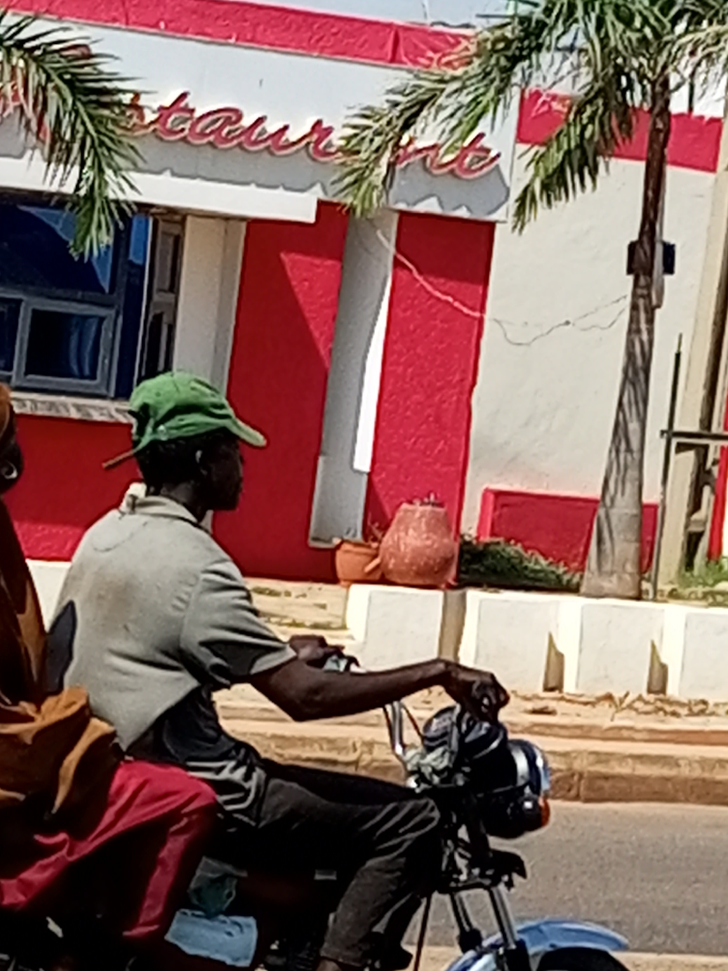 An outdoor, eye-level, medium shot captures a man wearing a green cap and a light grey polo shirt riding a motorcycle in the city of Tempure, Nigeria. He is seated with his left arm on the handlebars. Behind him, a person wearing a red and brown garment is visible. The motorcycle is facing to the right of the frame. In the background, a building with red and white stripes and palm trees can be seen. A sign with the word "restaurant" is mounted above a window on the building. The ground is paved.