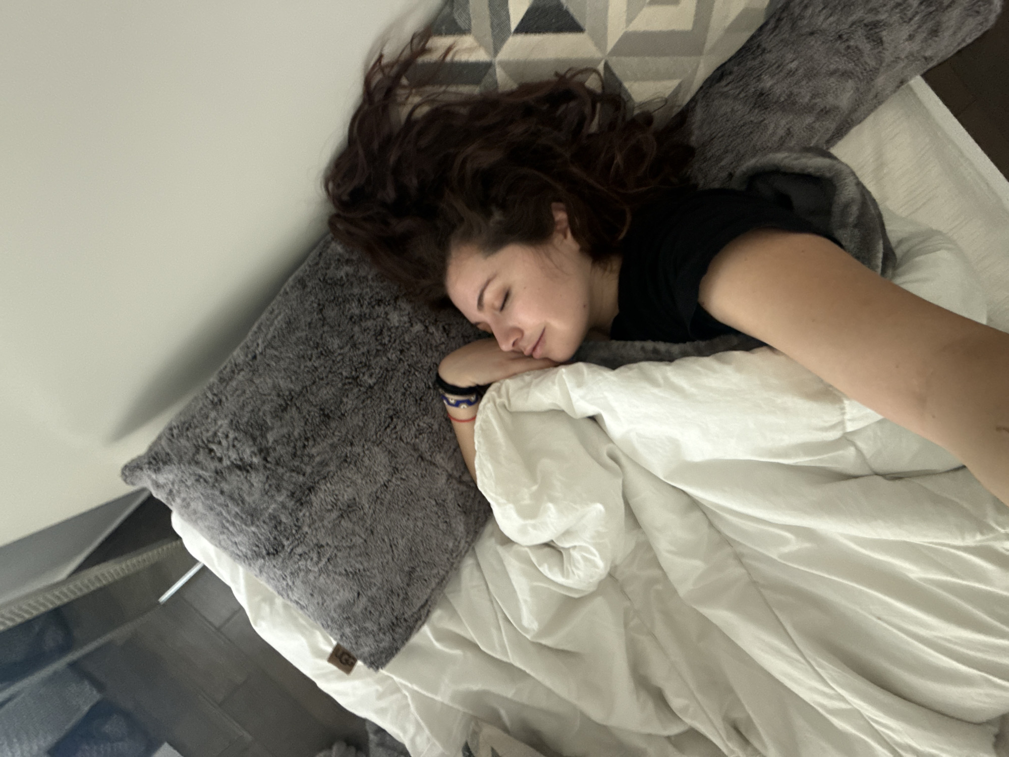 A young woman with long, dark, curly hair is sleeping on a bed. She is lying on her side, facing the camera. She is wearing a black T-shirt. Her left arm is bent and resting on a grey fluffy pillow with a wrist bracelet. The bed has white sheets and a grey furry pillow at the head. There is a patterned headboard. The room appears to be indoors. The location in Mexico City, Mexico is not discernible from the image.