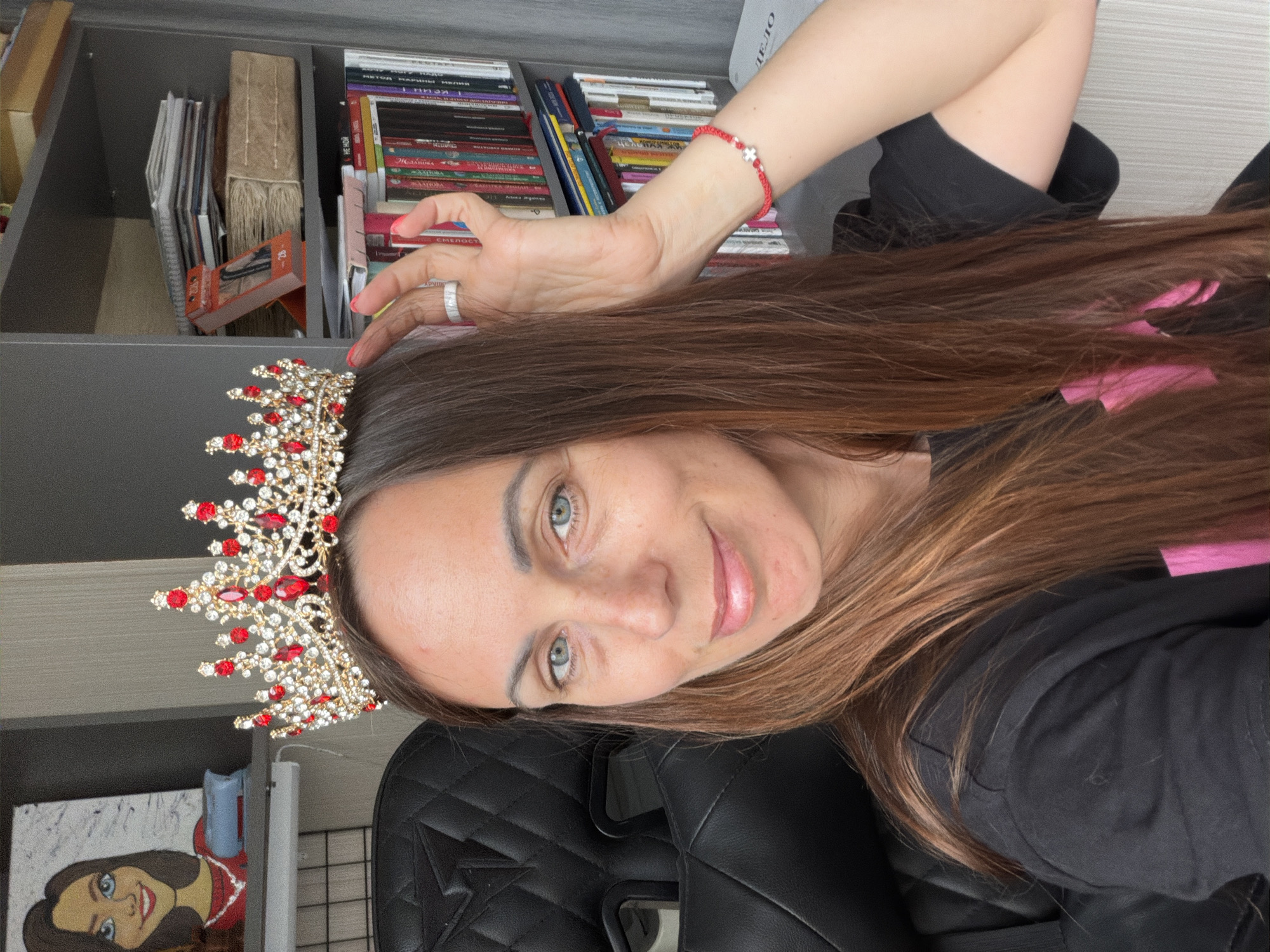 A woman with light skin and long brown hair is wearing a golden crown with red gems. She has light blue eyes and a light complexion. She is wearing a black shirt. She is sitting in a black leather chair. Behind her is a bookshelf filled with books and other items. There is a painting on the wall below the bookshelf. The location is undetermined, but the city mentioned is Birobidzhan, Russia.