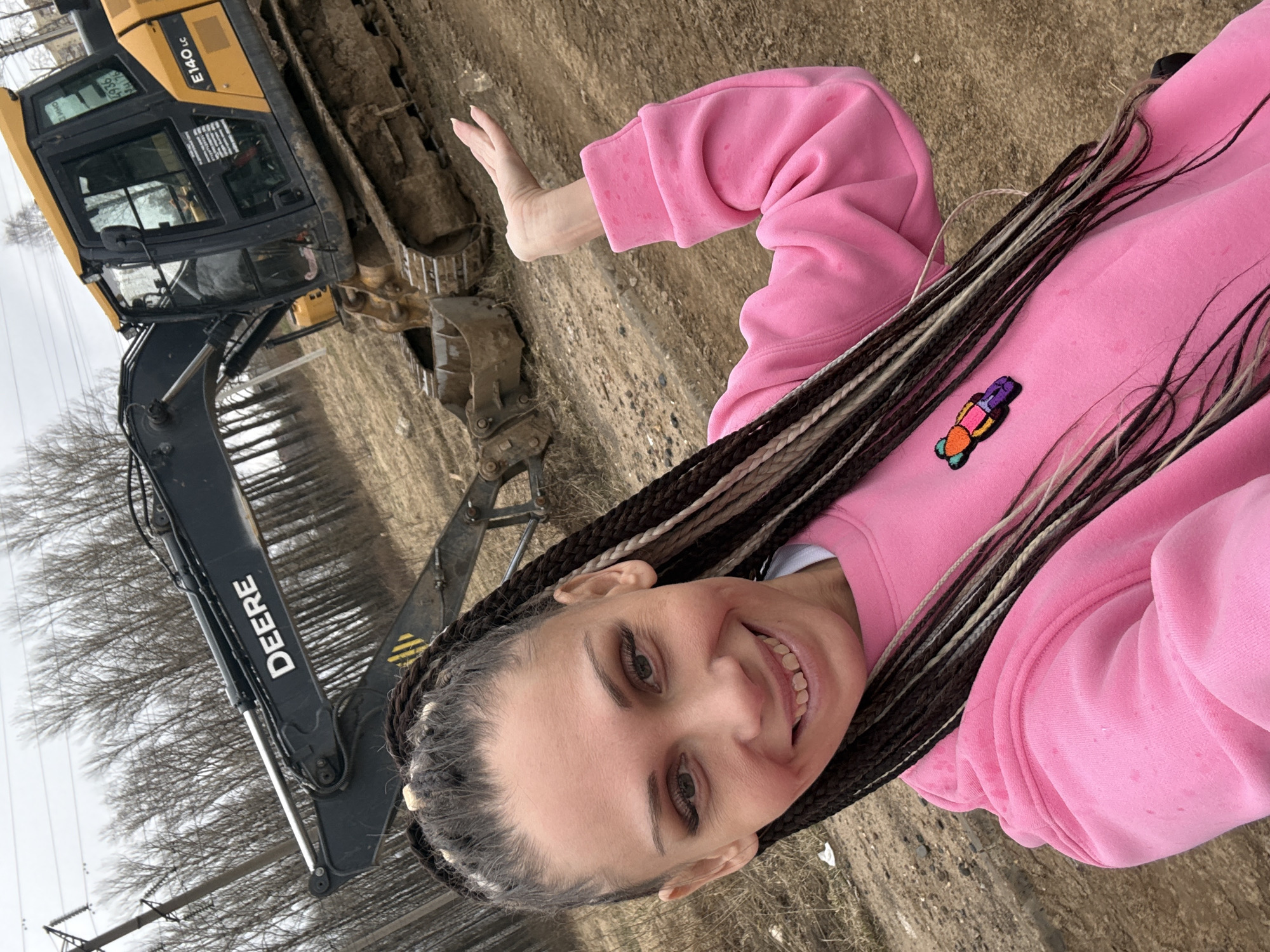 A smiling woman with long braids, wearing a pink sweatshirt with a colorful patch, is waving near a yellow and black John Deere E140LC excavator. The background features a dirt ground and trees. The location is Unknown, Unknown.