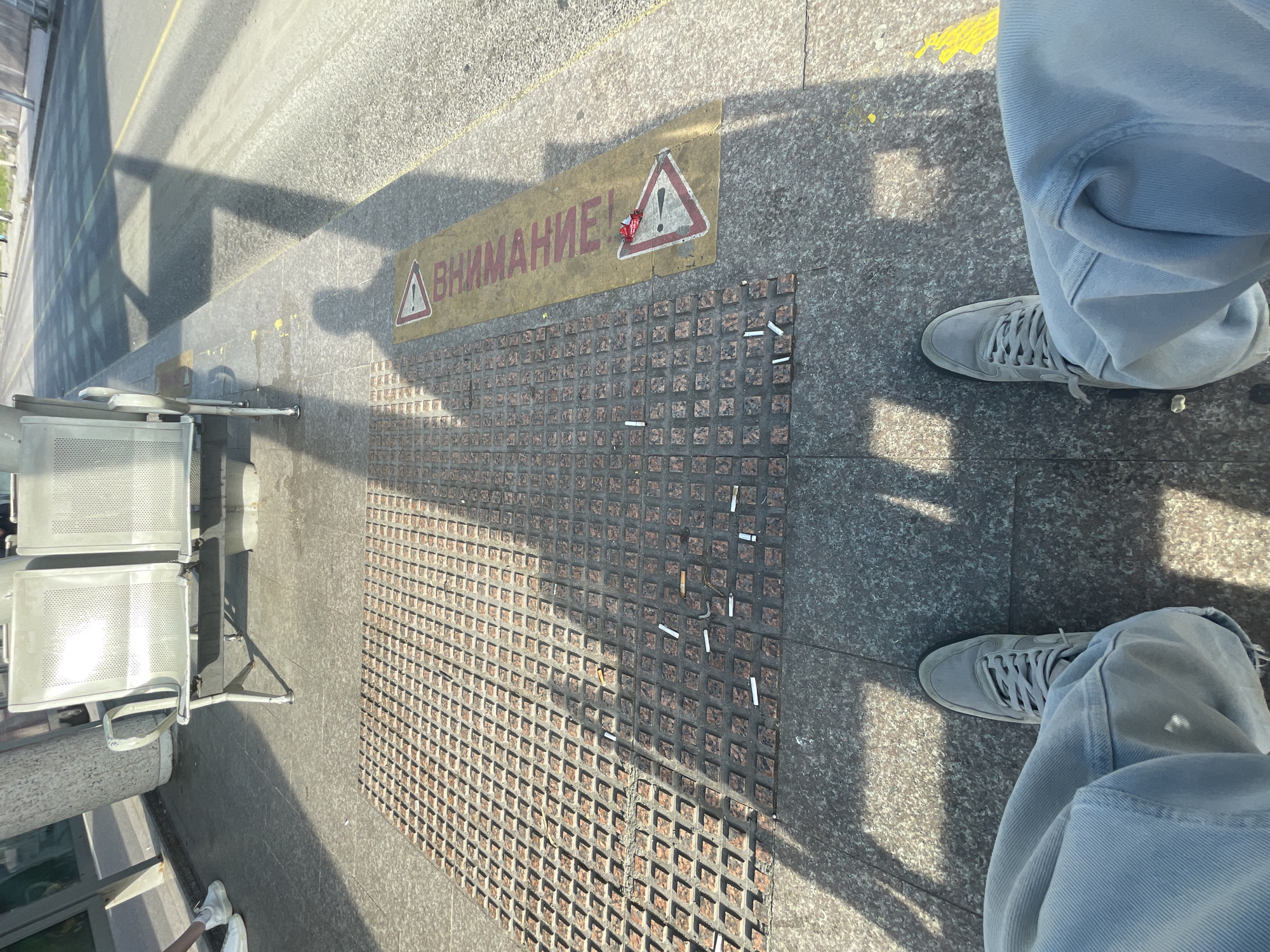 The image shows a section of a public space, likely a bus stop or train station in Burgas, Bulgaria. A sign in Russian, "Внимание!" (Attention!), with a caution symbol is visible, with some trash adhered to it. The ground is paved with patterned tiles, littered with cigarette butts. A pair of legs wearing light blue jeans and grey sneakers stands on the pavement. A row of metal chairs is positioned to the left. Sunlight casts sharp shadows across the scene.