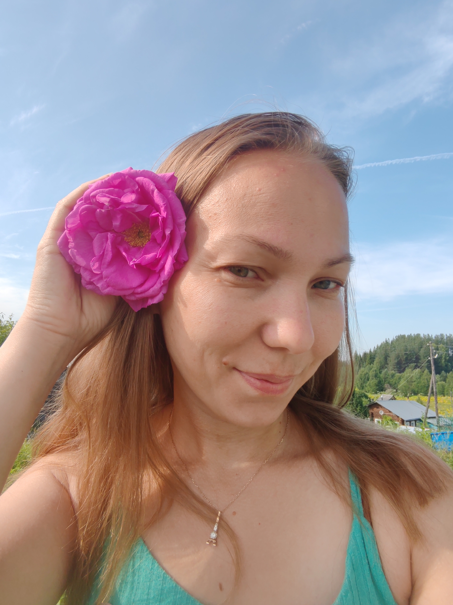 The image shows a selfie of a young woman with light brown hair, holding a pink rose behind her right ear. She is wearing a turquoise top and a gold necklace with a pendant. The background is a bright blue sky with some wispy clouds. The lower portion of the image shows a blurry view of a rural landscape with trees, houses, and yellow flowers. The woman is smiling and looking at the camera. The location is identified as Chukhlomsky District, Kostroma Oblast, Russia.