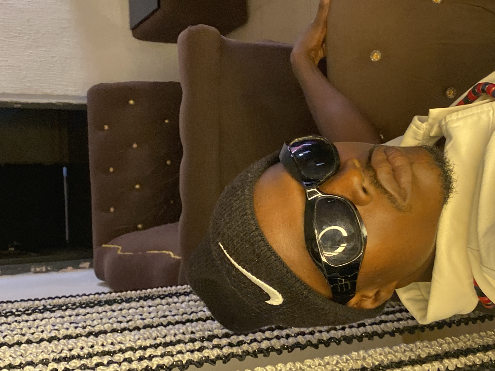 The image shows a close-up, waist-up view of a Black person wearing a black beanie with a white Nike logo, black wraparound sunglasses, and a light-colored hooded jacket. The person has a mustache and goatee and is reclining on a brown tufted sofa. A beaded curtain hangs to the left of the frame. The background consists of the sofa and a glimpse of a dark interior space, possibly a room in a residence or commercial establishment in Abakaliki, Nigeria. The lighting is dim, with the primary light source appearing to be from the front, casting shadows and highlighting the subject.