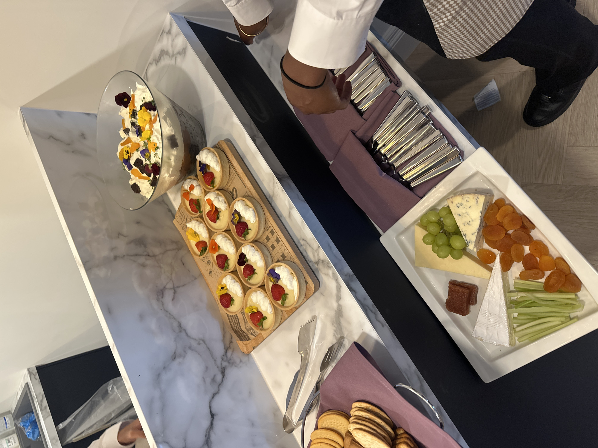 The scene depicts an indoor catering or buffet setup in Epsom and Ewell, United Kingdom. A marble-patterned countertop serves as a display surface, featuring a large glass trifle bowl filled with layers of dessert, topped with whipped cream, dark crumbs, and an assortment of yellow, purple, and reddish edible flowers. Adjacent to the trifle, a wooden board holds nine small tarts, each topped with white cream, a sliced strawberry, and small edible flowers. To the left, a set of silver tongs rests next to stacks of round crackers placed on a purple fabric napkin. Below this counter, a dark countertop provides a second display level. Here, a rectangular white platter showcases a variety of food items: a wedge of brie cheese, a slice of a light yellow cheese, a wedge of blue cheese, a cluster of green grapes, several dried apricot halves, and cut celery sticks. Another rectangular white tray contains multiple sets of silver cutlery (forks and spoons visible), each neatly rolled within a purple fabric napkin.
Two individuals are partially visible. On the left, a person wearing a light-colored long-sleeved shirt is seen reaching into an area behind the marble counter, with a clear plastic bag visible in the background. On the right, a person in a white long-sleeved shirt, black trousers, and a brown checkered garment (possibly an apron or lower part of a uniform) is standing next to the lower counter. Their right hand, adorned with a black hair tie on the wrist and a gold bracelet, is positioned above the cutlery tray. Their black dress shoe is visible on the light wooden floor beneath the counter.