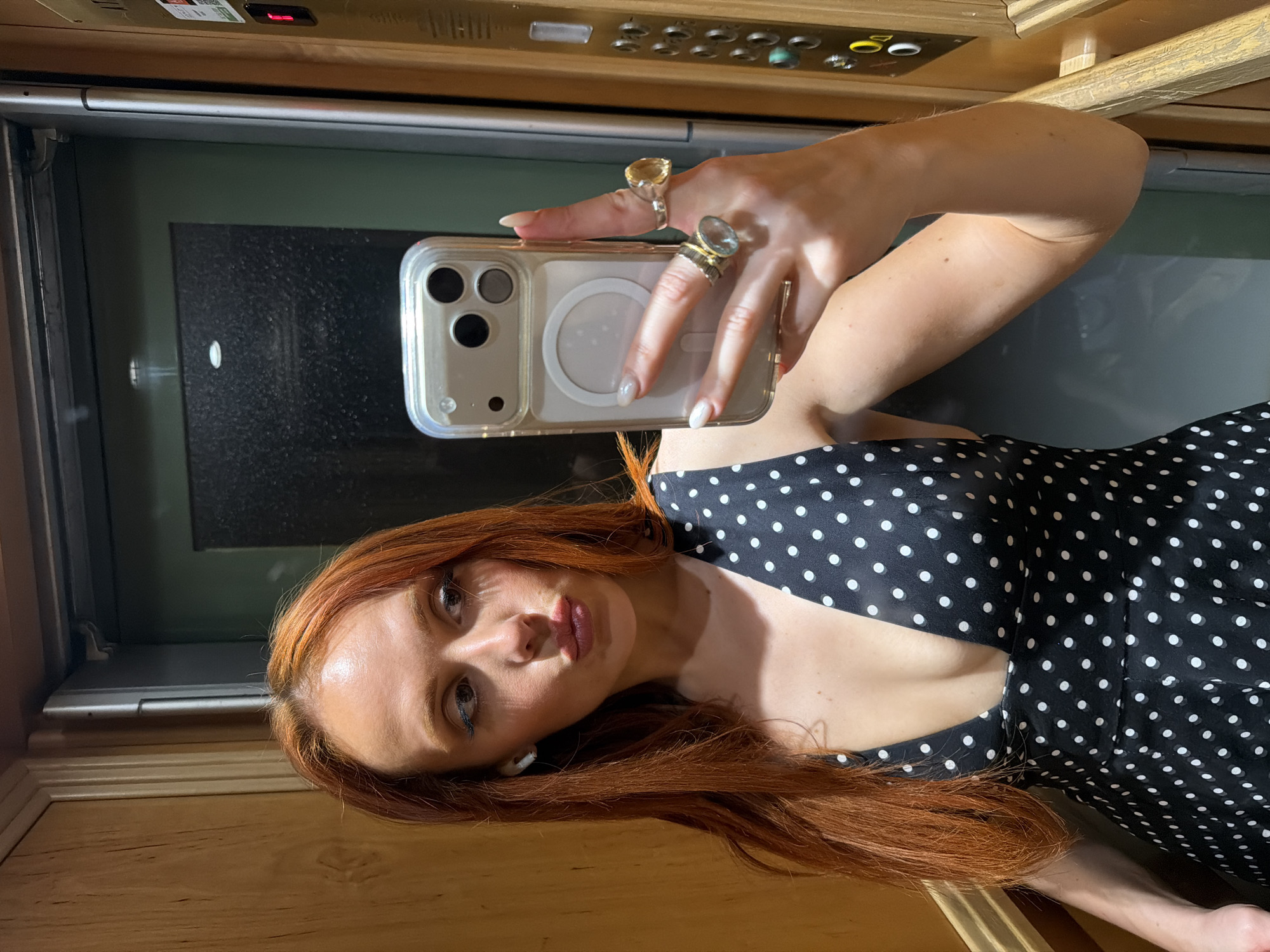 A reddish-haired woman is taking a selfie with a smartphone inside an elevator. She is wearing a black dress with white polka dots and has several rings on her fingers. The elevator has a wood-paneled interior and metal doors with a dark green-gray finish. The scene suggests the elevator is in a building in Paris, France.