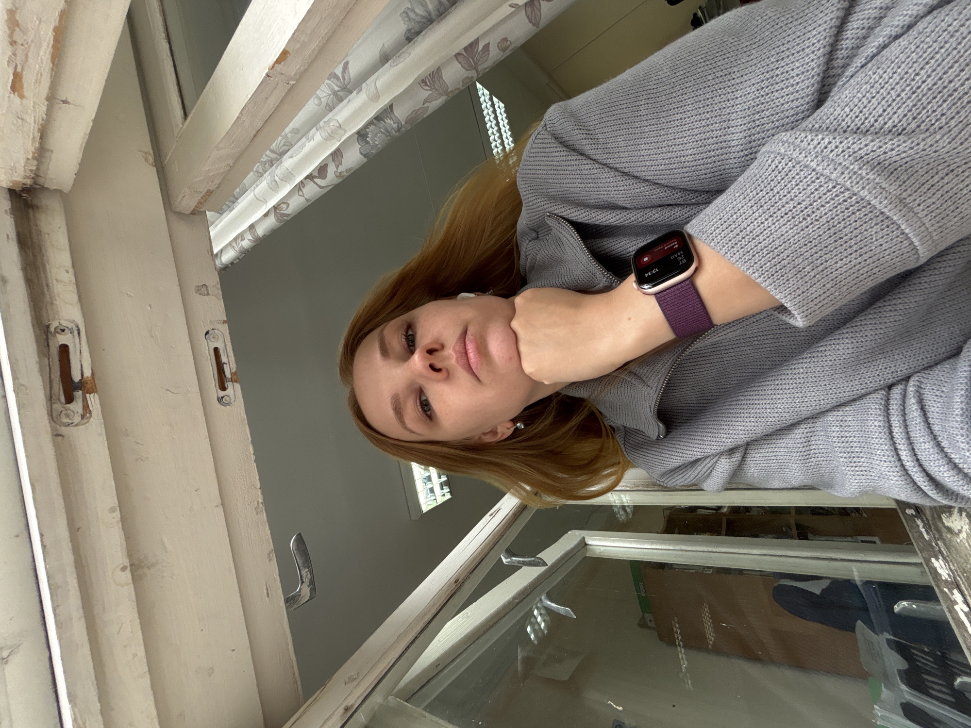 A woman with light brown hair, wearing a gray sweater and an Apple Watch with a purple band, rests her chin on her hand while looking out of an old, white-framed window. The window appears to be in a building in Rusanovo, Russia, based on the geotag. A floral-patterned curtain hangs partially in the upper left corner of the view. The window is partially open. The interior of the room visible through the window includes shelves with items.