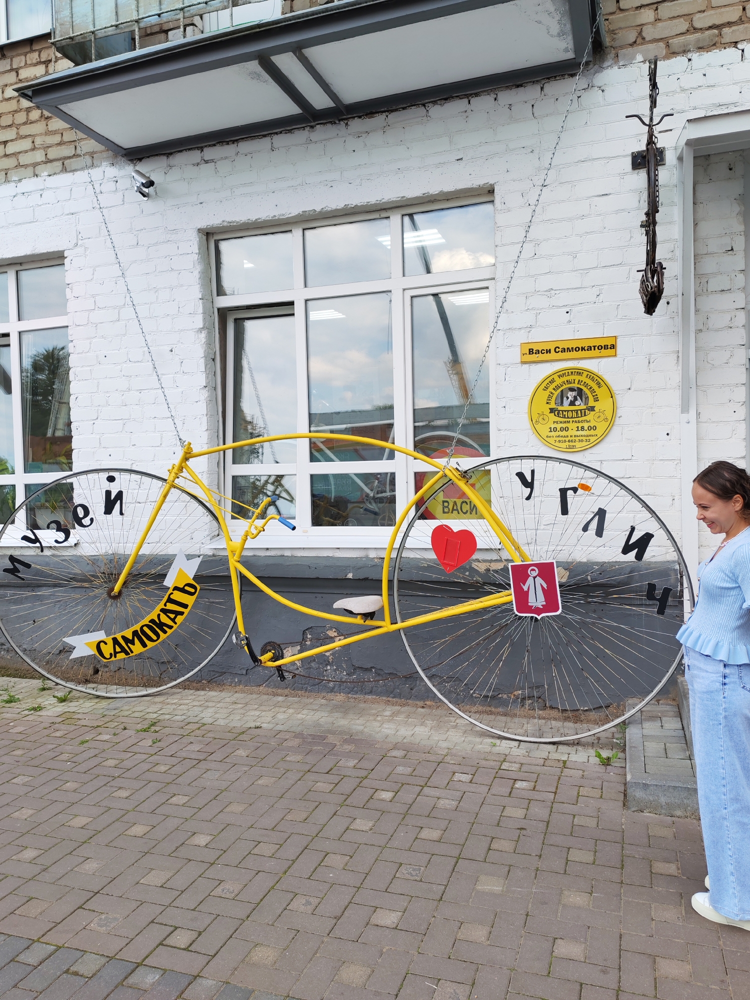 In the city of Selivanovo, Russia, a large, bright yellow bicycle sculpture is displayed on the facade of a white brick building. The bicycle is suspended by chains from above. Its frame and wheels are prominent, with the words "музей" (museum) written in black lettering on the left wheel and "самокатъ" (scooter) on a yellow banner attached to the left wheel. The right wheel has the Cyrillic letters "углич" (Uglich) in black. A red heart is affixed to the frame between the wheels, and below it, a red crest with a white figure is visible. A woman in a light blue top and jeans stands to the right of the sculpture, looking at it and smiling. A yellow sign with Cyrillic text, including "Ул. Васи Самокатова" (Vasya Samokatoa Street), is mounted on the building above the bicycle. A security camera is also attached to the building to the left of the sculpture. The building has large windows and is situated on a paved walkway.
