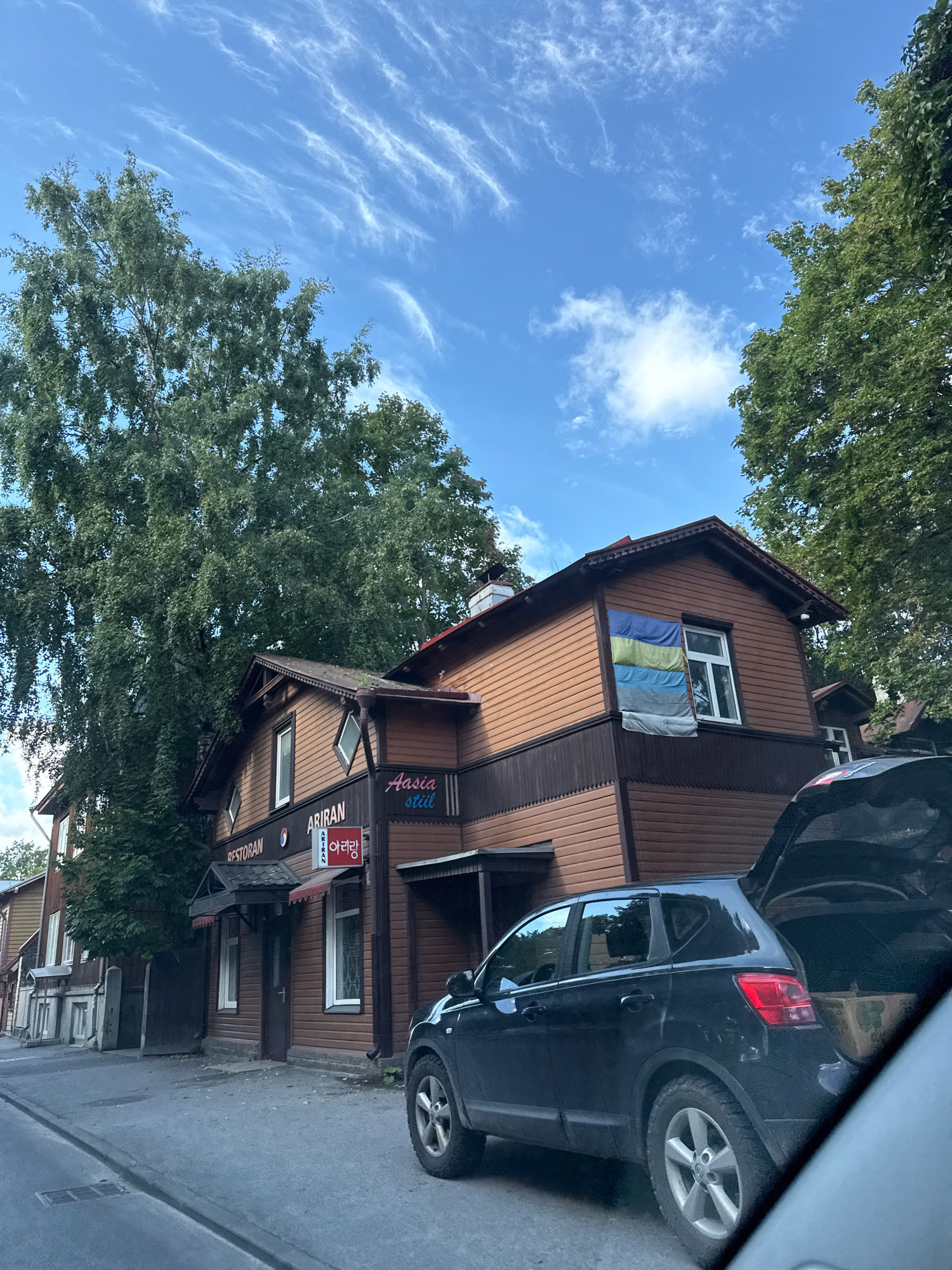 The image depicts a street view in Tallinn, Estonia, specifically near the "Ariran" restaurant. A two-story wooden building, painted in shades of brown, is prominent. The restaurant's signage, "RESTORAN" and "ARIRAN," is visible. The upper floor of the building has a window displaying a flag composed of horizontal stripes of blue, yellow, and gray. To the right of the building, a black SUV is parked with its trunk open, revealing a cardboard box inside. The street is paved and a sidewalk is present. Large trees with green foliage line the street, and the sky is blue with wispy white clouds. The overall scene appears to be a daytime setting.