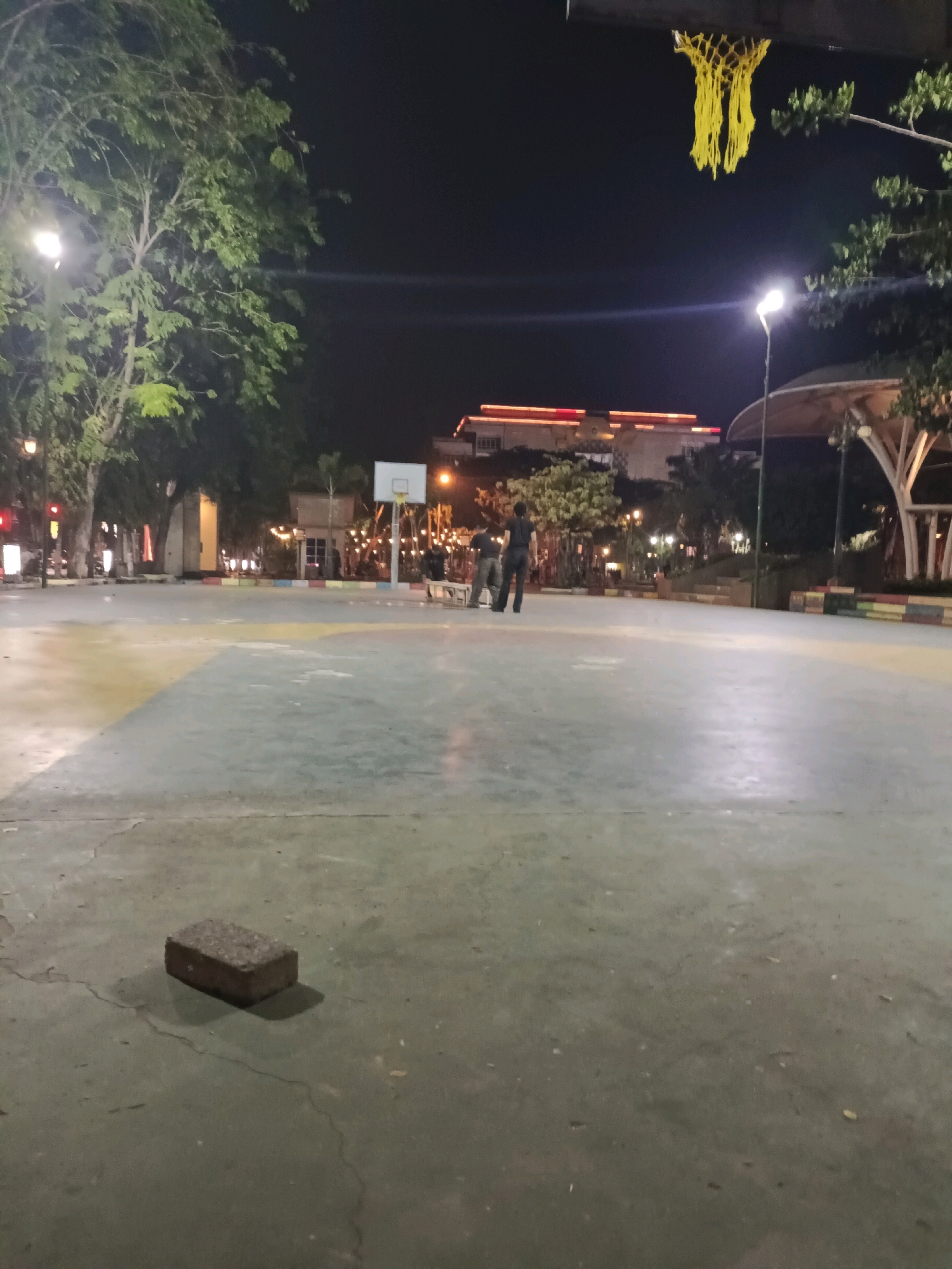 The image depicts an outdoor basketball court at night in Lamongan, Indonesia. The court surface is concrete with colored markings, and a basketball hoop with a yellow net is visible. Two individuals are standing near the hoop, one appearing to be interacting with equipment, while the other stands nearby. Trees with green foliage line the perimeter of the court. In the background, buildings with illuminated windows and decorative lights are present. Streetlights and other artificial lighting illuminate the scene. A single brick lies on the court in the foreground. The overall impression is of a public recreational space after dark.