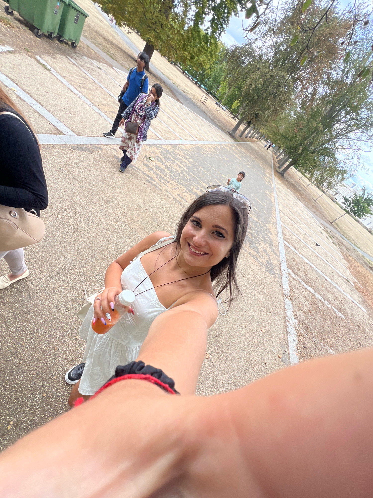 A wide-angle, low-light selfie captures a woman in the foreground holding a bottle of orange liquid. She is smiling and wearing a white, sleeveless dress. A red and black scrunchie is visible on her left arm. In the mid-ground, other individuals are walking on a paved path lined with trees. To the left, two large green bins are visible. The background shows a park-like setting with more trees and buildings. The overall scene suggests an outdoor activity in a park in London, likely during daytime.