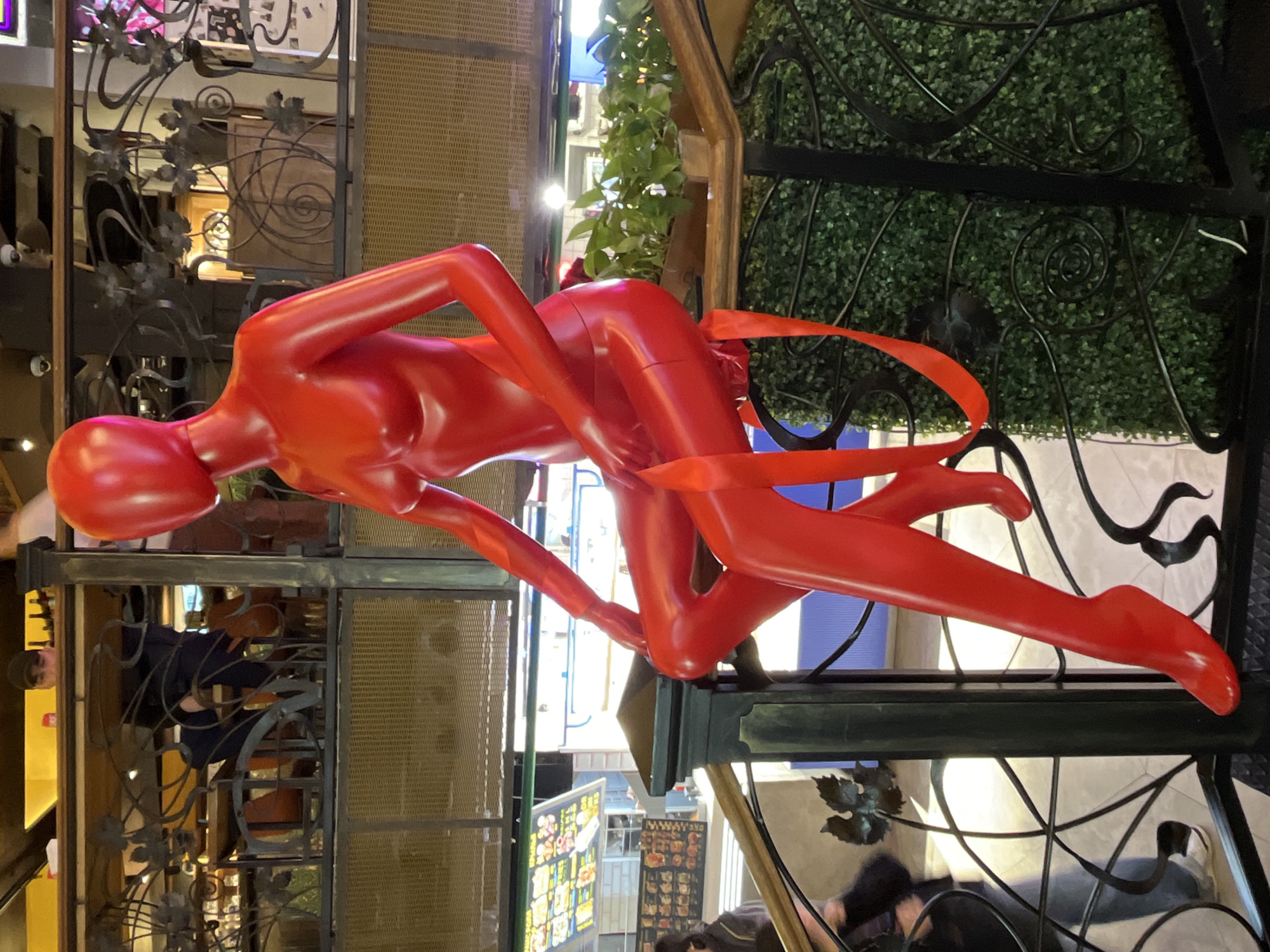 The image captures an art installation within a public space, likely a shopping mall or commercial center, in Moscow, Russia. The central element is a bright red, abstract sculpture of two intertwined human figures. The sculpture is placed atop a decorative wrought iron structure adorned with artificial greenery. A red ribbon is draped loosely around the sculpture. The surrounding environment includes other retail spaces, metal latticework, and a partially visible security camera. Several people are present in the background, engaged in activities like shopping or using electronic devices. The overall lighting is bright and diffuse.