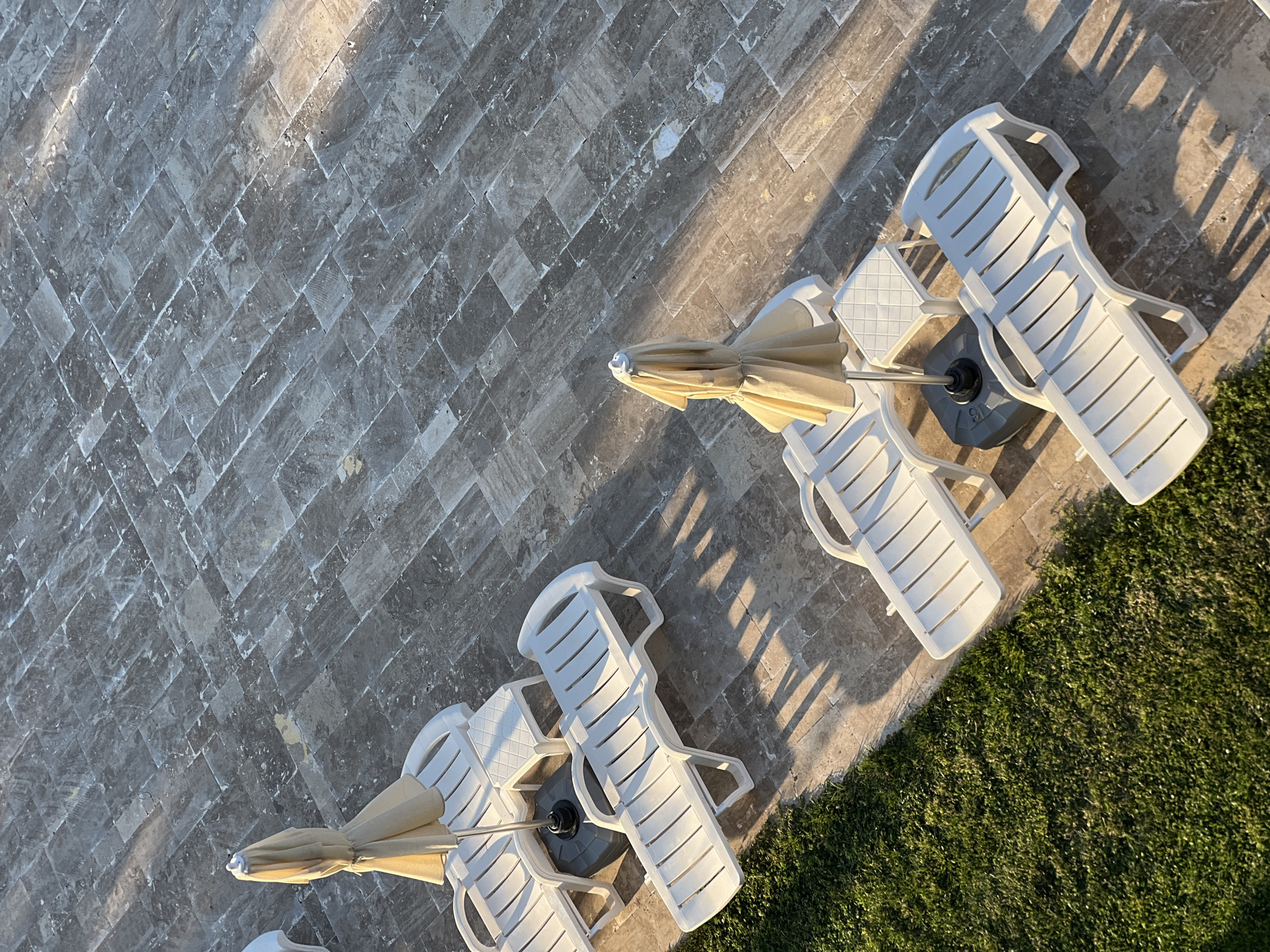 The image depicts an outdoor scene, likely a poolside or patio area, with several white plastic lounge chairs and umbrellas. The ground is paved with large, rectangular grey tiles laid in a diagonal pattern. To the left of the tiled area is a strip of vibrant green grass.
In the foreground, there are three lounge chairs. The closest one on the right is empty and positioned diagonally across the frame. To its left and slightly behind it, another lounge chair is also empty, with its shadow cast across the tiles. Further back and to the left, a third lounge chair is partially visible, also empty. Two of the lounge chairs have folded umbrellas standing next to them, one pale yellow and the other a similar light color. A small white side table is situated between two of the lounge chairs in the middle ground.
The scene is illuminated by what appears to be low-angle sunlight, creating distinct shadows from the lounge chairs and umbrellas that stretch across the tiled surface. The lighting suggests it is either early morning or late afternoon. There are no people visible in the image.
The location is identified as being in the city of Yeşilova, Turkey.