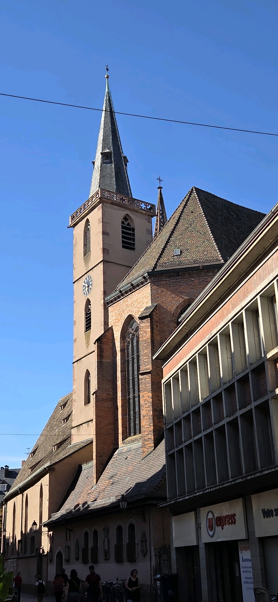 The image shows a street view in Strasbourg, France, with a prominent church tower and spire dominating the left side. The church has a tall, rectangular tower made of light-colored stone, featuring arched windows and a clock face. Above the tower, a pointed spire covered in dark grey material rises into a clear blue sky. The church also has a steeply pitched roof covered in terracotta tiles. Adjacent to the church, on the right, is a modern building with a facade of vertical concrete elements and glass panels.
In the foreground, the street is paved, and several people are visible walking or standing. Bicycles are parked near the buildings. The atmosphere appears to be daytime, with sunlight casting shadows on the buildings. The scene suggests a typical urban environment with a mix of historical and modern architecture.