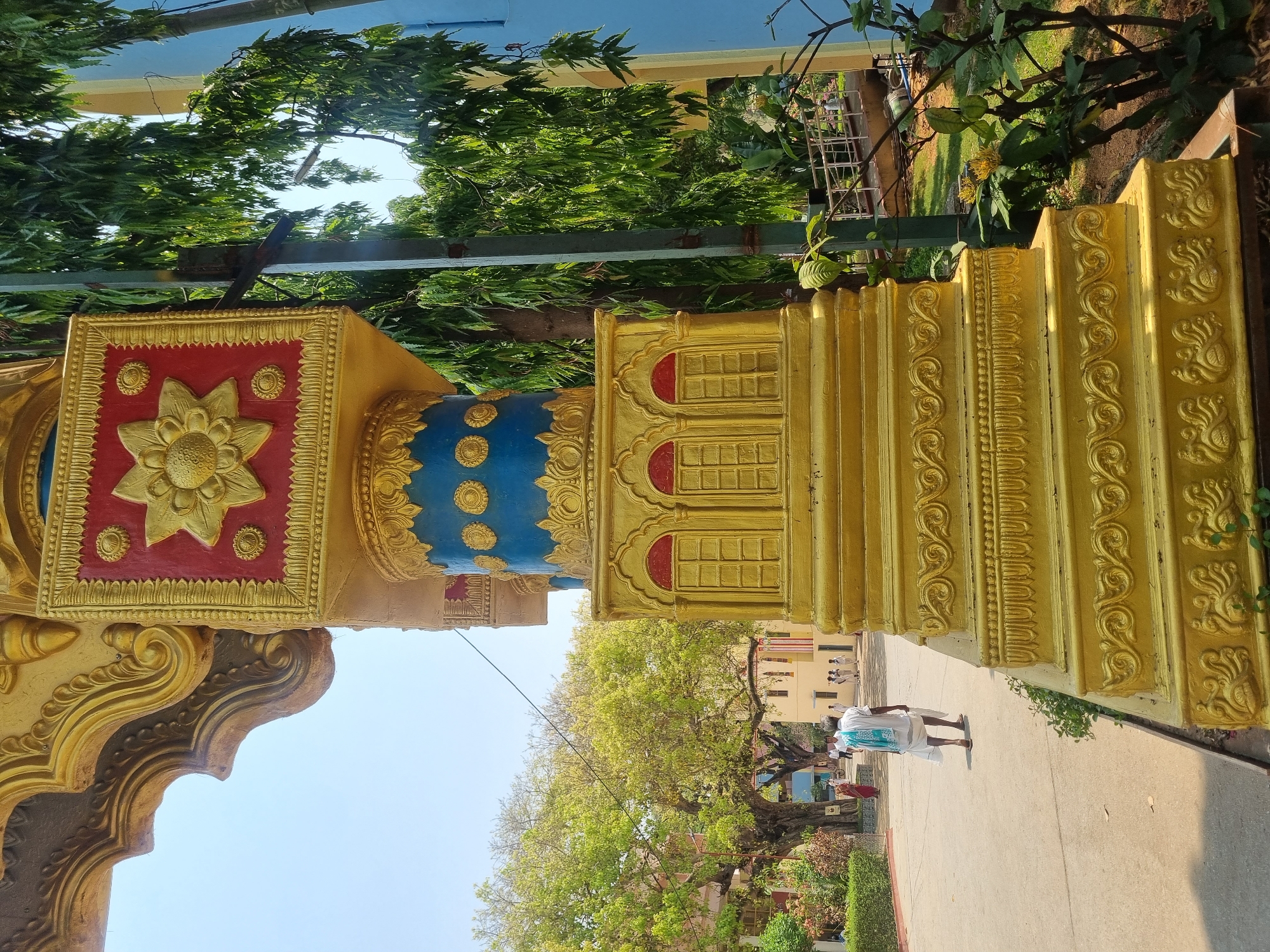 The image captures an outdoor scene in Puttaparthi, India, within a large architectural complex, likely an ashram or temple courtyard, under clear daylight.
In the foreground, occupying the right side of the frame, is a prominent, ornate golden pillar. Its upper visible portion features a square red panel centered with a detailed gold sunburst or floral motif, surrounded by gold geometric borders. Below this, a section of the pillar is blue with recurring gold circular designs. The lower section of the pillar is golden, presenting multiple tiers of carved ornamentation including three arched, recessed "window" designs with red interiors and grid patterns. The pillar's base is stepped, adorned with intricate scrollwork and stylized, possibly zoomorphic, carvings.
To the left and center, a wide, light-colored paved pathway or courtyard extends into the background. A single man, viewed from behind, walks away from the camera on this path. He wears a white dhoti and a light blue and white striped or patterned shawl draped over his left shoulder, appearing to wear sandals.
In the background, to the left, a large, mature tree with a dense canopy of bright green leaves provides shade. Beyond this tree, a light-colored building is partially visible. To the right background, behind the golden pillar, dense, slender-leafed green trees are visible alongside a vertical utility pole. A light blue building facade is also present on the far right. Several other individuals are visible in the far distance on the paved area, some in white attire, and one person appears to wear a red sari. The sky is clear and light blue, indicating bright, sunny weather with discernible shadows.