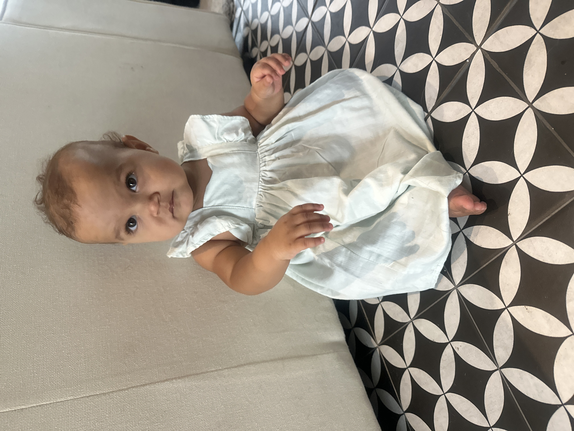 A baby wearing a white dress is lying on a white sofa that is next to a black and white patterned floor. The baby is facing the camera, with their head on the sofa and their feet on the floor. The photo was taken indoors. No information regarding a specific location in North Kuta, Indonesia, is provided in the image.