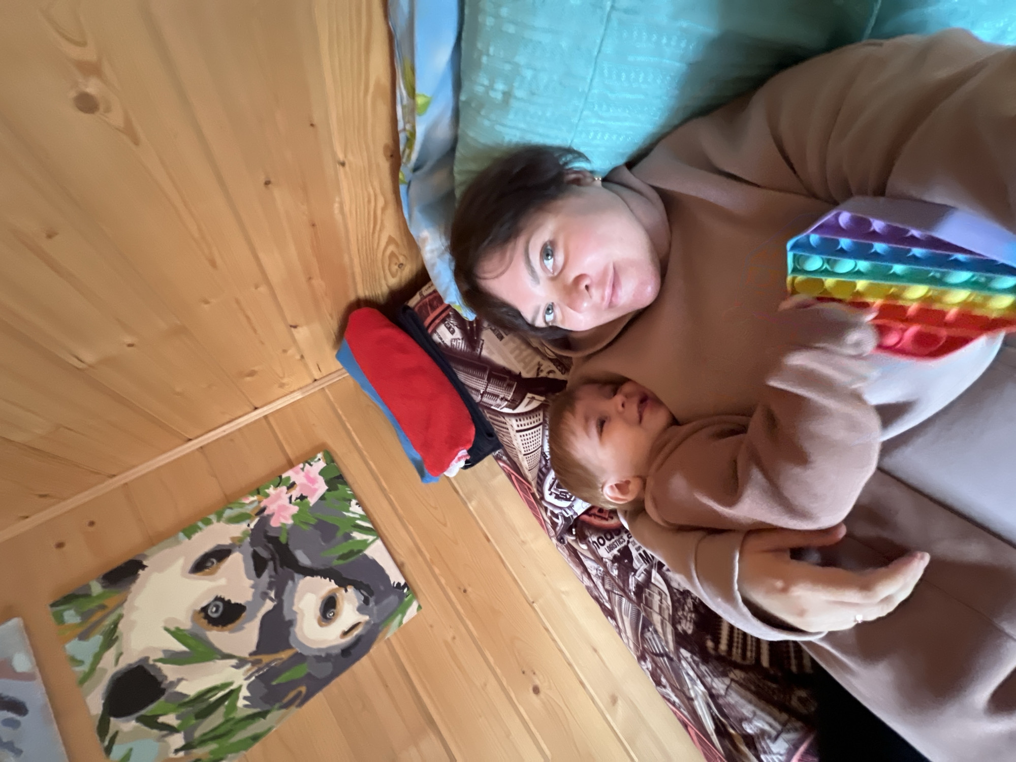 A woman and a baby lie on a bed. The woman is looking at the camera and holding a rainbow-colored pop-it toy. The baby is nestled in her arms. The bed is covered in patterned linens. A painting of a panda bear hangs on the wooden wall. The location is possibly in Protasy, Russia.