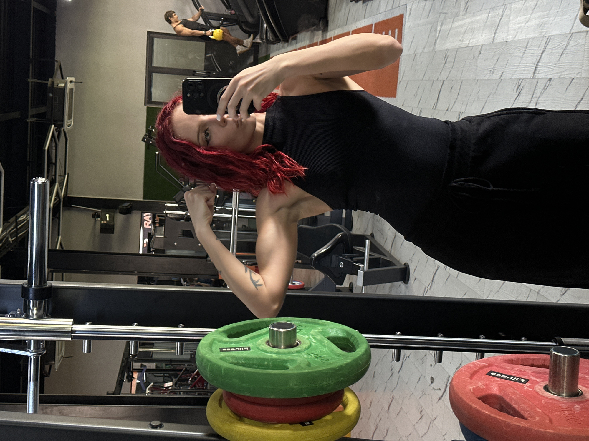 The image depicts an indoor gym scene in North Kuta, Indonesia. In the foreground, a female with red, wavy hair, dressed in a black ribbed sleeveless top and black sweatpants, is captured taking a mirror selfie with a black smartphone held in her right hand. She is posing with her left arm flexed, revealing a bicep and a tattoo on her upper arm. To her immediate left, part of a chrome Smith machine is visible, loaded with a stack of weight plates: yellow, red, and a green 10KG plate labeled "ESENFIT." Another red "FITNESS" branded weight plate is visible lower on the machine. The gym floor is composed of light-toned, patterned tiles, featuring an orange and white lane marking stripe. In the background, visible through the mirror's reflection, a male individual with dark hair and a muscular physique, wearing a black tank top and light-colored shorts, is operating a stair climber machine while holding a yellow kettlebell in his right hand. The gym environment includes light grey walls, dark ceiling structures with lighting, and additional gym equipment partially visible.