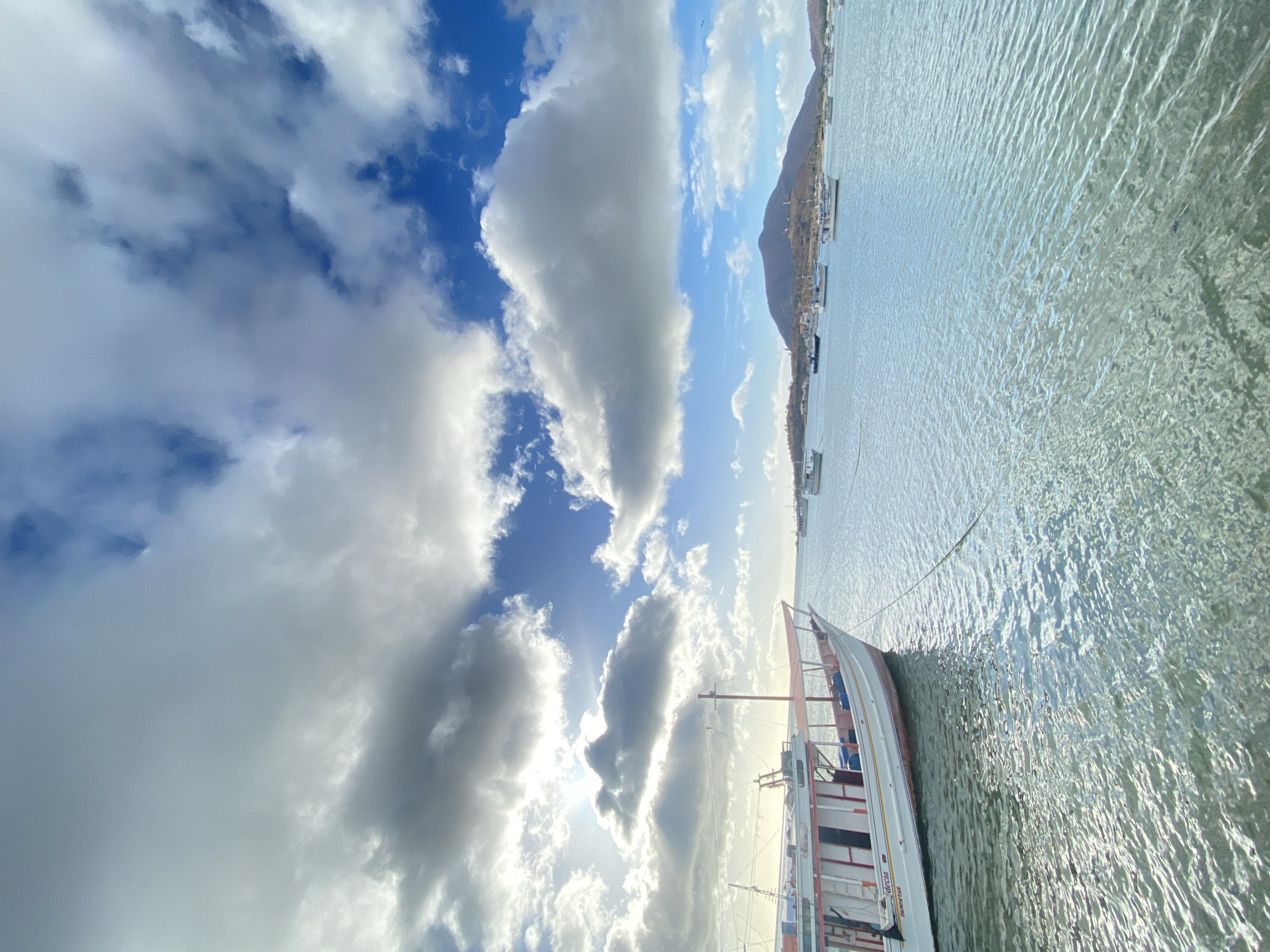 The image shows a partially visible boat on the water with a mountainous shoreline in the distance. The sky is filled with clouds and patches of blue. The location appears to be in Juan Griego, Venezuela, as indicated by the shoreline and the style of the boats. The water shows ripples and reflections.
