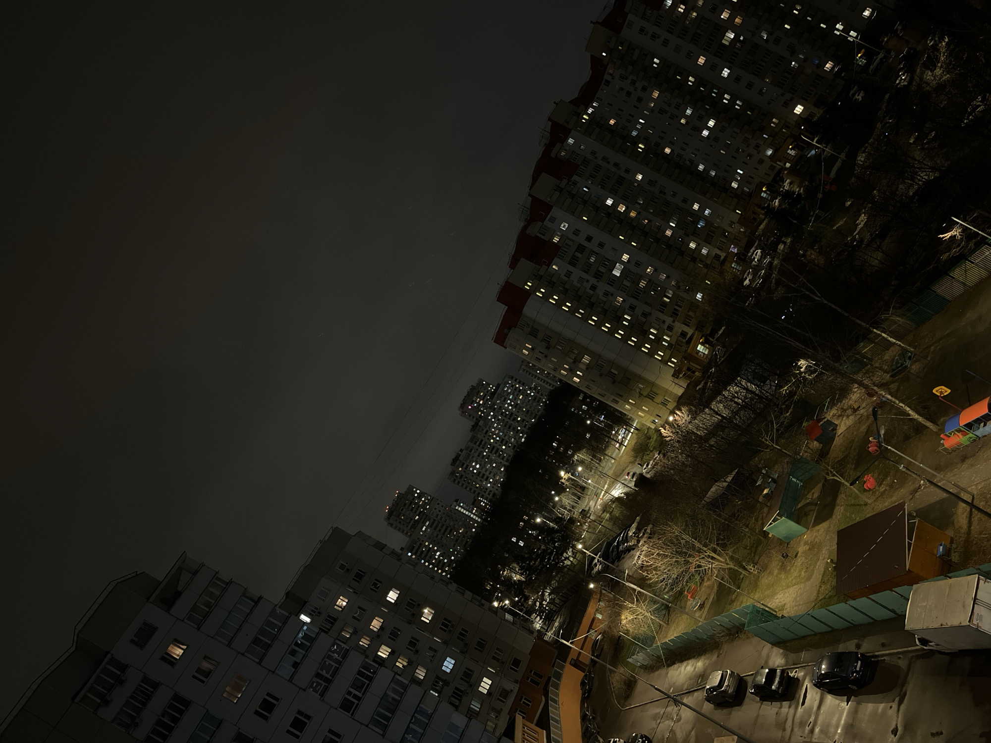 The image depicts an urban scene at night, likely in a densely populated area of Moscow, Russia. The composition is dominated by multi-story apartment buildings with numerous illuminated windows, suggesting residential occupancy. In the foreground, a courtyard area is visible, featuring trees, possibly a playground, parked cars, and storage units. The sky is overcast. No people are directly visible in the image.