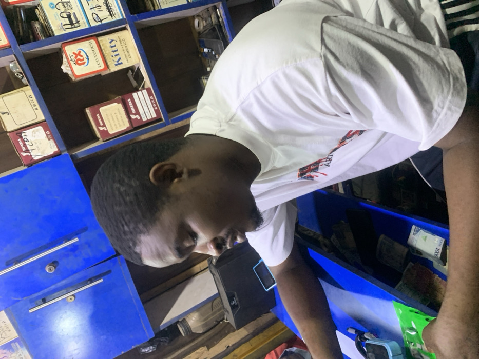 An indoor scene, likely a shop or business in Garaku, Nigeria, features a man positioned centrally, looking downwards to his right. The image is taken at an oblique angle, making the man and the environment appear tilted. He is a dark-skinned individual with short, dark hair and a neatly trimmed beard or goatee, wearing a white short-sleeved t-shirt with some red and black graphic designs on the front.
Behind the man, a wall of blue-painted wooden shelves is visible, filled with numerous boxes and items, suggesting an inventory of goods. Some boxes have discernible labels, including "KITTY," "LCD CAMBO LCD," and "TOUCH," along with a red and white circular logo displaying "LCB Touch." Below these shelves, there are blue cabinet-like structures with drawers or compartments.
To the man's lower right, on a counter or shelf, stacks of Nigerian currency are clearly visible, with "1000" printed on the top notes, indicating 1000-naira denominations. Beside the money, a black electronic device, possibly a smartphone, rests. Further to the right, a light blue container or box with a handle is partially visible. The lighting is artificial, illuminating the foreground and parts of the shelves, while some areas are darker. The overall context suggests a retail or service environment, possibly dealing with electronics or phone parts, where the man is engaged in a transaction or task.