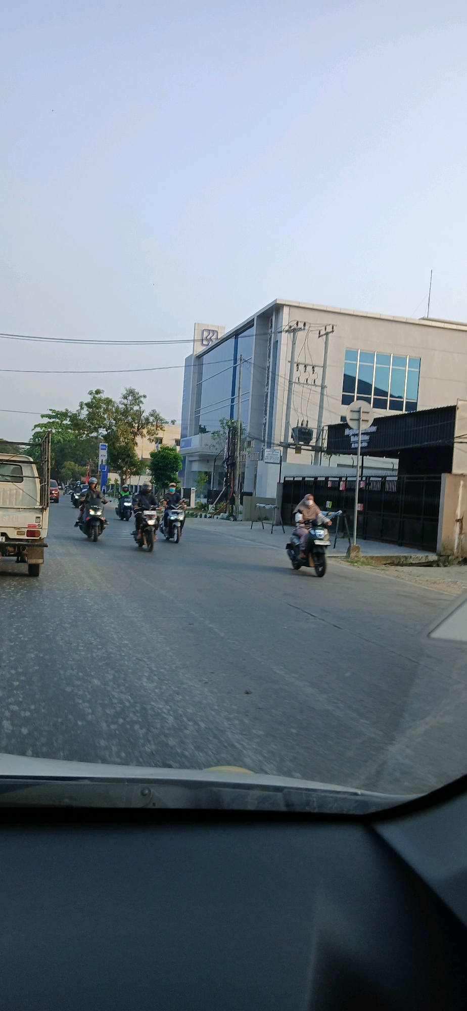 The image captures a street scene in Samarinda, Indonesia, with a modern building in the background. The building, which appears to be an office or commercial establishment, features a glass facade and a distinct logo that resembles two curved lines forming an abstract shape, possibly a bank or corporate logo. Power lines and poles are visible along the street, indicating urban infrastructure.
The road is asphalt, showing signs of wear and some scattered debris. Several motorcycles are present, with riders dressed in casual clothing, some wearing helmets. One motorcycle in the foreground carries a rider wearing a hijab. A white truck is parked on the left side of the road. The sky is clear and pale blue, suggesting it is either early morning or late afternoon. The overall impression is a typical day in a bustling Indonesian city.