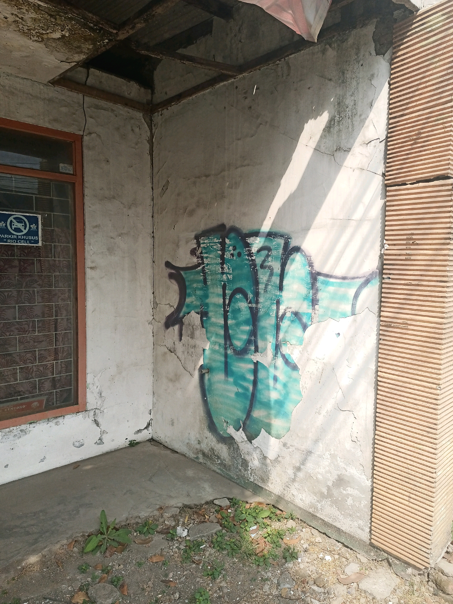 The image shows the corner of a dilapidated building in Malang, Indonesia. The building has a concrete wall with significant cracks and peeling paint, a damaged roof structure with exposed beams, and a window with an orange frame. Graffiti in shades of turquoise and purple covers a portion of the wall. Adjacent to the wall is a fluted pillar made of beige material. On the ground in front of the building, there is a concrete floor covered with scattered debris, dry leaves, and small plants. A blue sign with white text and an icon depicting a crossed-out car is visible on the window.