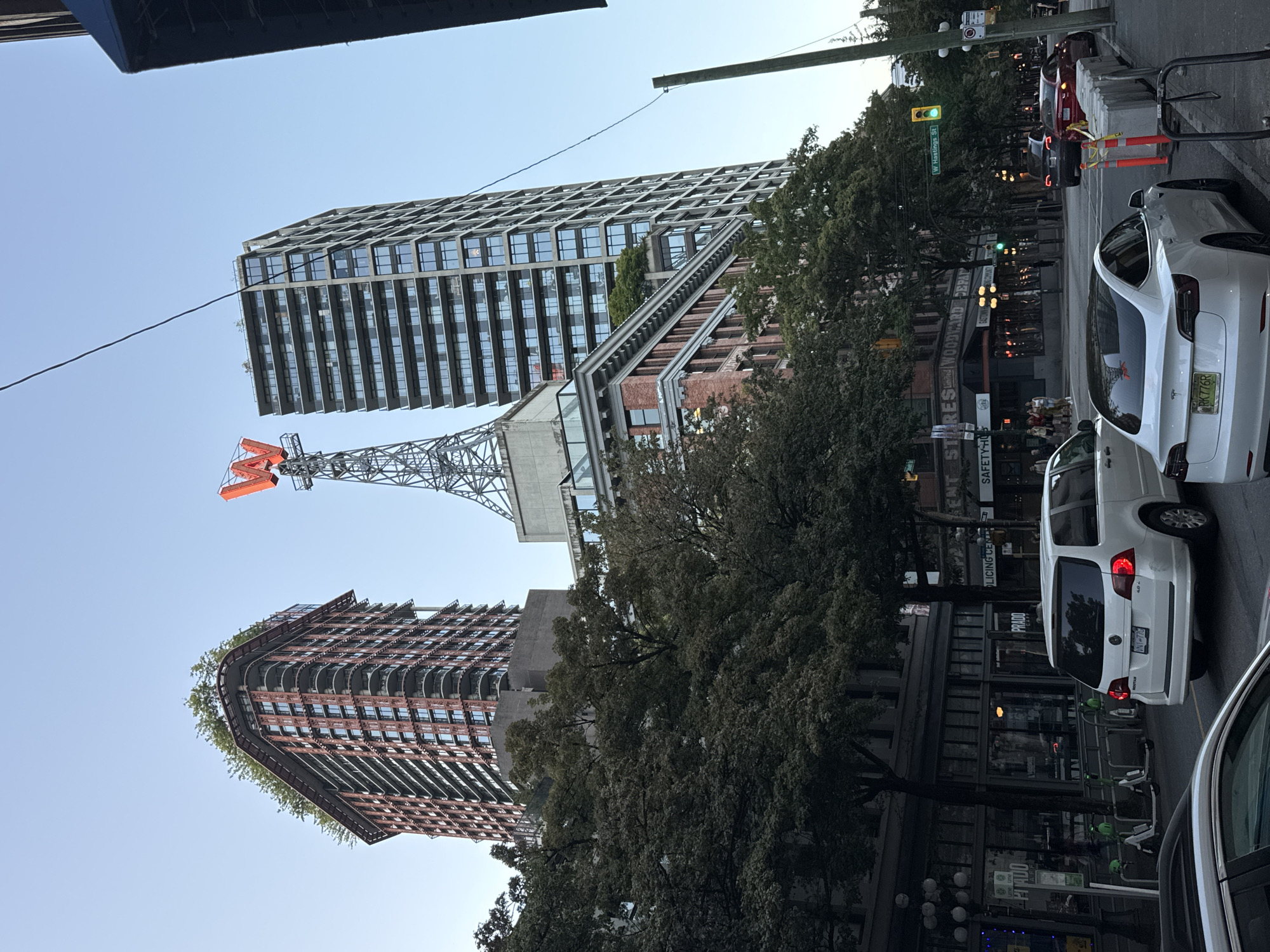 This is a street-level, slightly low-angle shot of a cityscape in Vancouver, Canada. Dominating the scene are two tall, modern buildings. The building on the left is a mixed-use high-rise with a reddish-brown facade and balconies, topped with a green roof garden. Adjacent to it, and slightly behind, is a sleek, dark gray residential tower with numerous balconies. Between these two prominent structures, an old broadcast tower with a large, orange "W" logo at its apex is visible. Below these buildings, older brick structures with storefronts line the street.
The street itself is paved and appears to be a busy urban thoroughfare. Several vehicles are present, including a white SUV, a white Tesla, and a red sedan. The white Tesla in the foreground has a license plate that reads "RK776R". Pedestrians are visible on the sidewalk, walking past storefronts. Trees with dense foliage line the sidewalk, casting shadows. Traffic lights are visible, with one showing green. Street signs indicate "W Hastings St". The sky above is clear and a pale blue, suggesting either early morning or late afternoon. The overall atmosphere is that of a vibrant, active urban environment in downtown Vancouver.