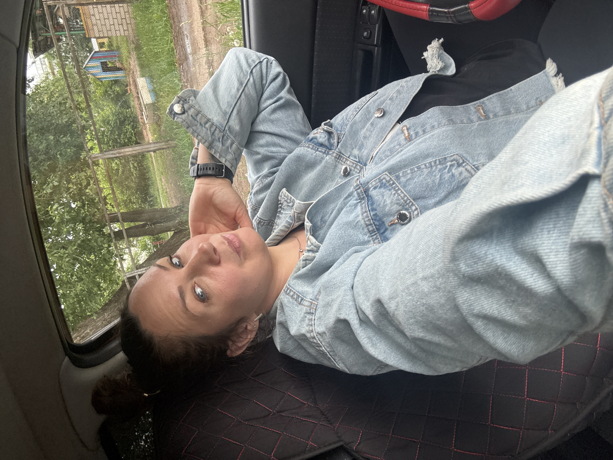 A female individual, estimated to be in her 30s-40s, is seated in the driver's position of a vehicle, taking a selfie. Her dark brown hair is pulled back into a bun. She wears a light-wash denim jacket with an unfinished hem over a black top, and black trousers are partially visible. A black digital watch is worn on her left wrist, and a small silver-colored stud earring is visible in her left ear. Her left arm is bent, with her hand supporting her head near her jawline. The car's interior features a black quilted seat cover with red stitching, a black door panel with four window control buttons, and a partial view of a black steering wheel with a red accent on the right.
Through the car window, an outdoor environment in Chaikovsky, Russia, is visible. The scene features a green, unkempt grassy area with mature trees. A wire mesh fence with metal posts runs horizontally across the mid-ground. Beyond the fence, a small, multi-colored wooden structure resembling a playhouse, painted with vertical stripes of blue, green, red, and yellow, is present. To its right, a low, white and blue trough-like object and a small brick structure are visible. The ground immediately beyond the fence appears to be a dirt path or unpaved area with damp patches. Further in the background, more trees and a distant, light-colored building are partially obscured.