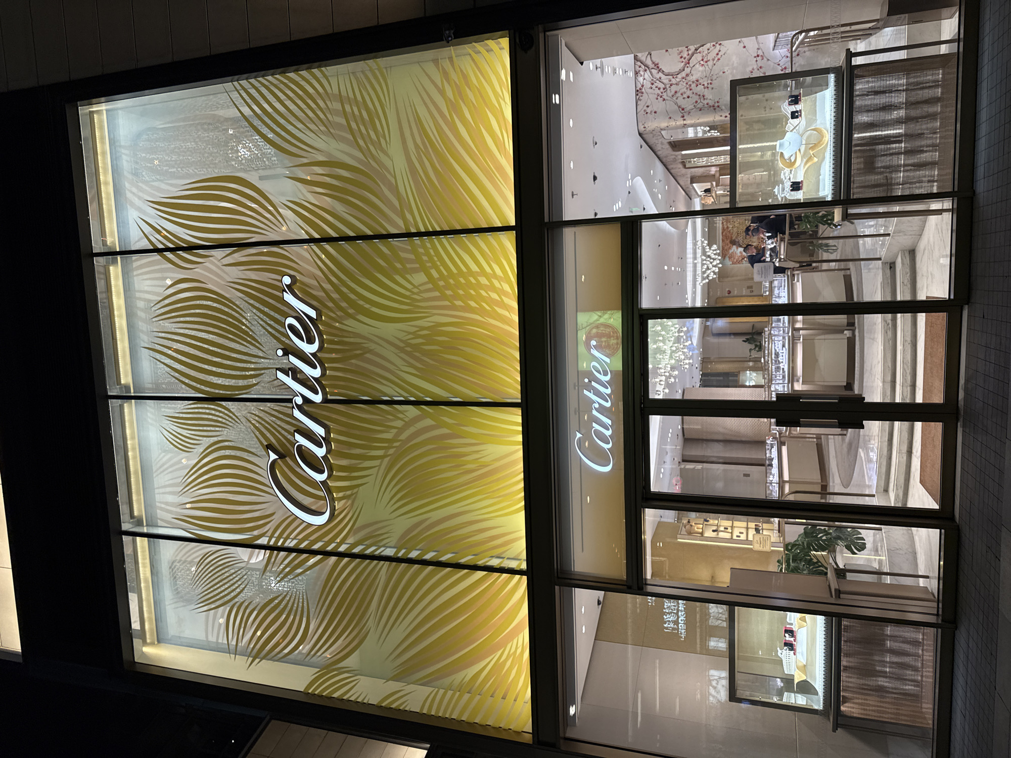 Here is a description of the image:
The photograph depicts the exterior of a Cartier store at night. The store's front features large windows with the Cartier logo prominently displayed. The windows on the left have a decorative yellow and white patterned design. Through the glass, the store's interior is visible, revealing multiple levels, display cases showcasing jewelry, interior decorations, and a small number of people. The environment outside the store appears urban, with minimal street visibility. The image does not confirm that it is in the Dongcheng District, China.
