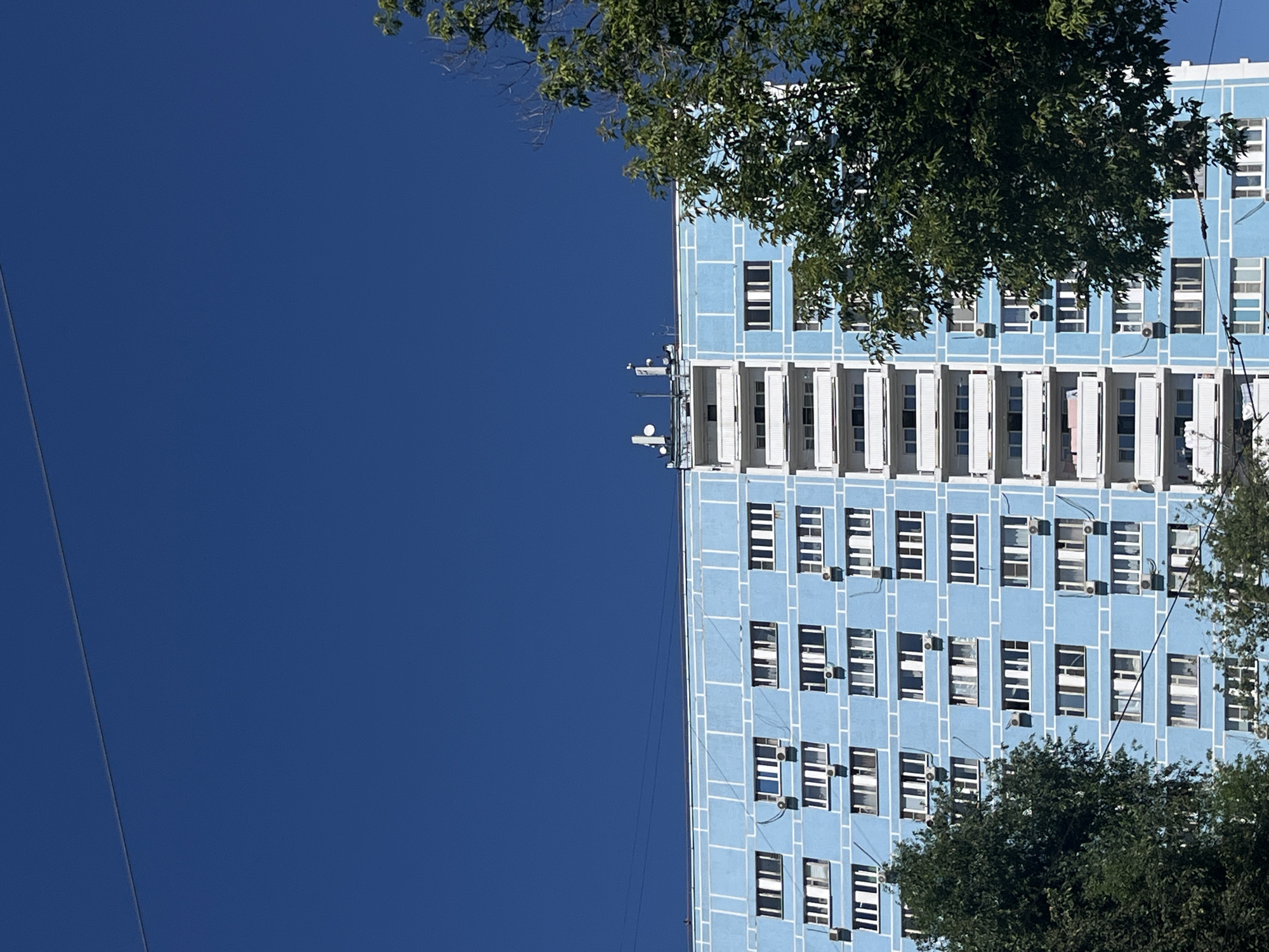 A low-angle outdoor shot shows a light blue multi-story apartment building against a clear, dark blue sky. The building has a symmetrical facade with numerous windows and balconies. The windows are white-framed and appear to be of various sizes. Some windows have air conditioning units attached. The balconies are white with railings. On the roof of the building, there are several antennas and what appear to be communication equipment. Trees with dark green foliage partially obscure the sides of the building. Power lines are visible running horizontally across the top of the frame. The image appears to be taken in a residential area of Moscow, Russia, given the architectural style of the building and the presence of power lines. No people are visible in the image.