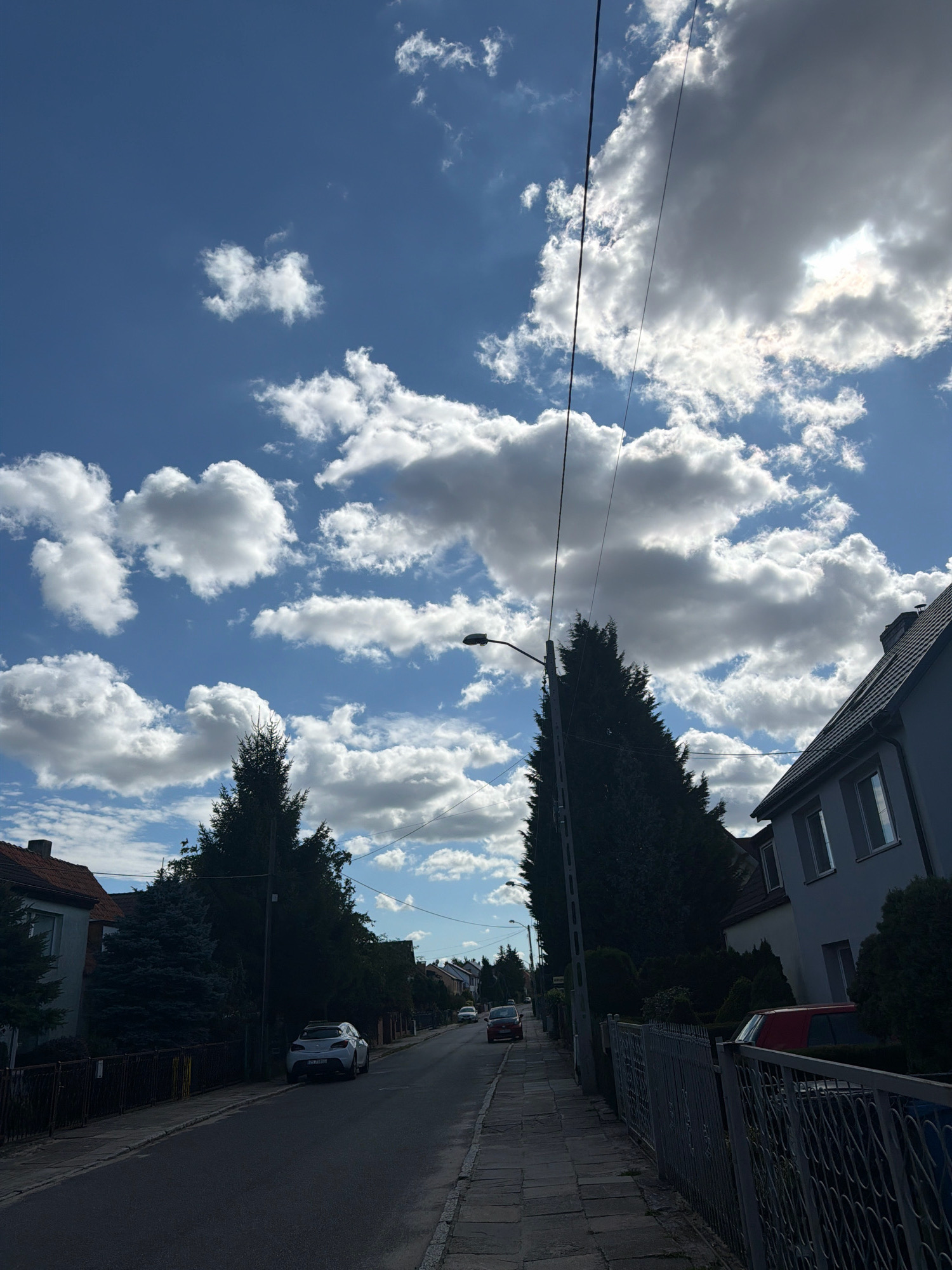 The image captures a street scene in Szczecin, Poland, characterized by a bright blue sky filled with fluffy white and grey clouds. The sun is partially obscured by a large, dark cloud on the right side of the sky. A streetlamp stands on a pole to the left of a tall, dark evergreen tree. On the right side of the street, a light-colored residential building with gray roofing and visible windows is situated. A dark metal fence runs along the sidewalk. On the left side of the street, houses with red-tiled roofs are visible, along with a dark fence. A white car is parked on the side of the road, and the street itself is paved with asphalt. Further down the street, more cars are visible, along with more residential buildings and trees. Overhead power lines are also present in the scene.