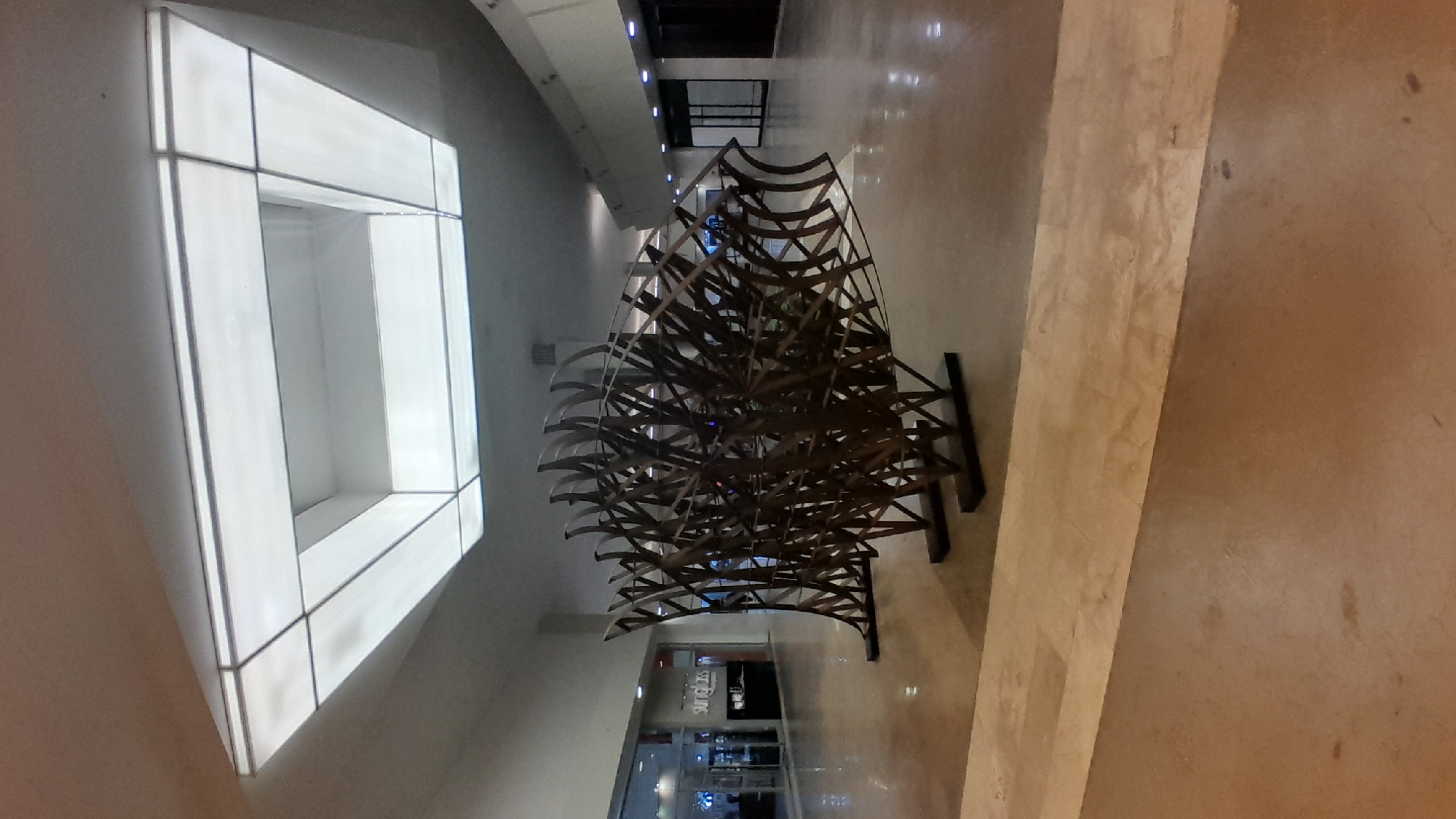 In Pampatar, Venezuela, inside a brightly lit mall, a large, abstract metal sculpture resembling a fish is prominently displayed. The sculpture, made of interconnected, curved metal pieces, stands on a dark base. In the background, stores with glass storefronts are visible, one of which has a sign that reads "sunglasses." The mall floor is polished and reflective, with a lighter-colored strip running horizontally in the foreground. The ceiling features a large, rectangular light fixture. No people are visible in the immediate vicinity of the sculpture.