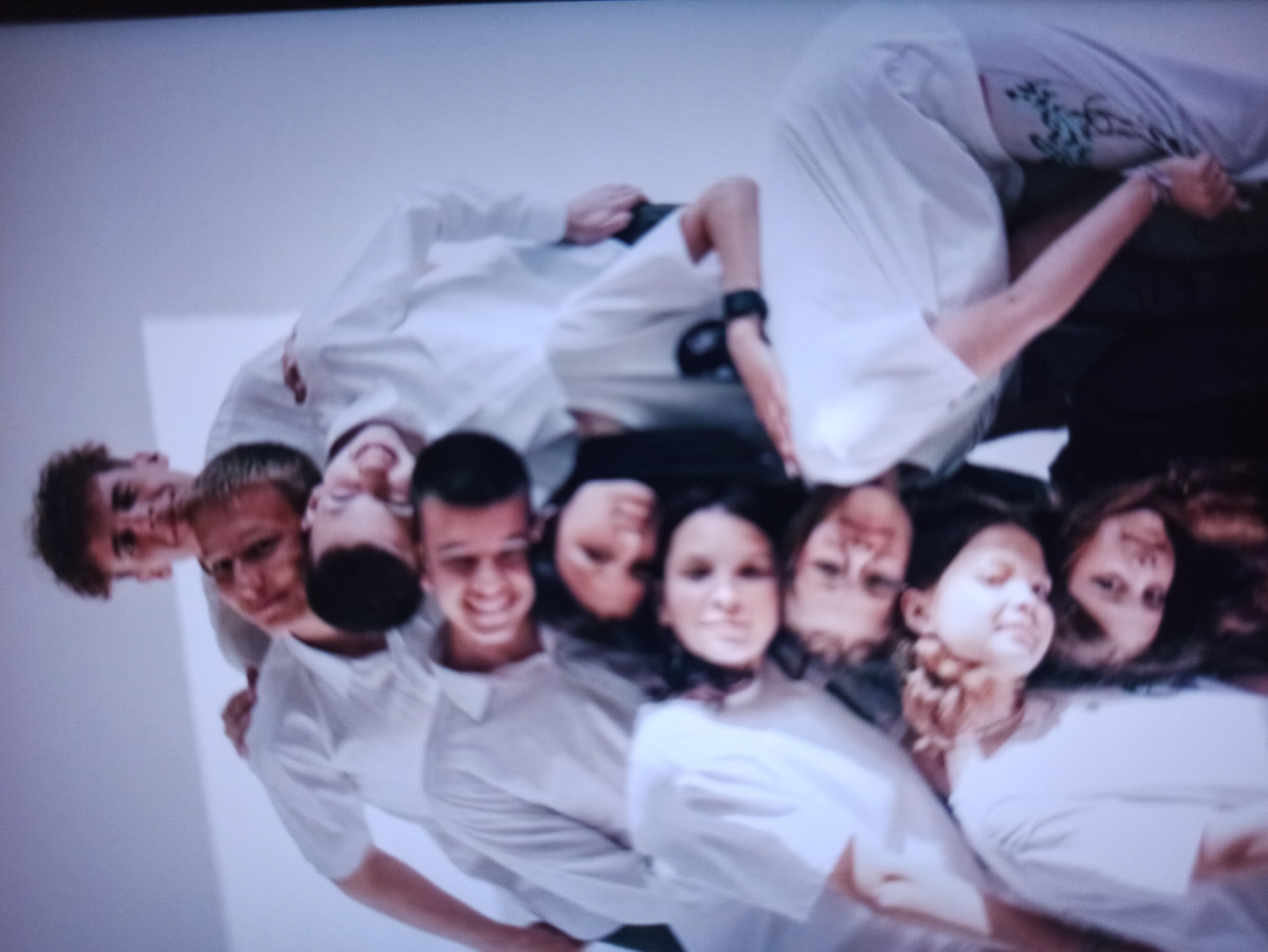 A group of nine young individuals, appearing to be in their late teens or early twenties, are arranged in a cascading, layered pose against a plain white background. They are all wearing loose-fitting white tops, possibly shirts or sweaters, and some are wearing matching light-colored pants with floral tattoos visible on one of the individuals. The individuals are stacked on top of each other, with their heads and shoulders forming a human pyramid. The person at the very top is looking forward with a slight smile, while the subsequent individuals are positioned at various angles, some with their eyes closed, some looking down, and some looking towards the camera. The overall impression is one of a posed, artistic photograph, likely taken in a studio setting.