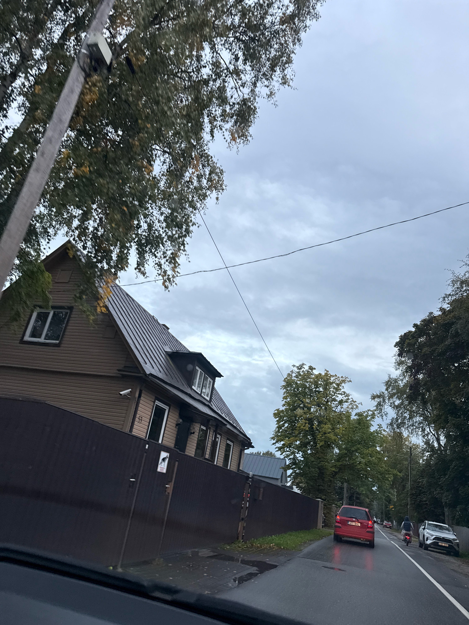 The image depicts a cloudy day in Tallinn, Estonia, as viewed from a car. In the foreground, the edge of a car's dashboard is visible. A dark brown fence runs along the side of a street. Behind the fence is a two-story wooden house with a dark metal roof. The house has several windows, some with white frames. A security camera is mounted on the side of the house. The street is wet, indicating recent rain, and a red car is driving away from the camera. Further down the street, a person on a bicycle is passing a white SUV. Trees line the street, and their leaves suggest it is autumn. Overhead, telephone wires are strung across the sky. The overall atmosphere is overcast and somber.