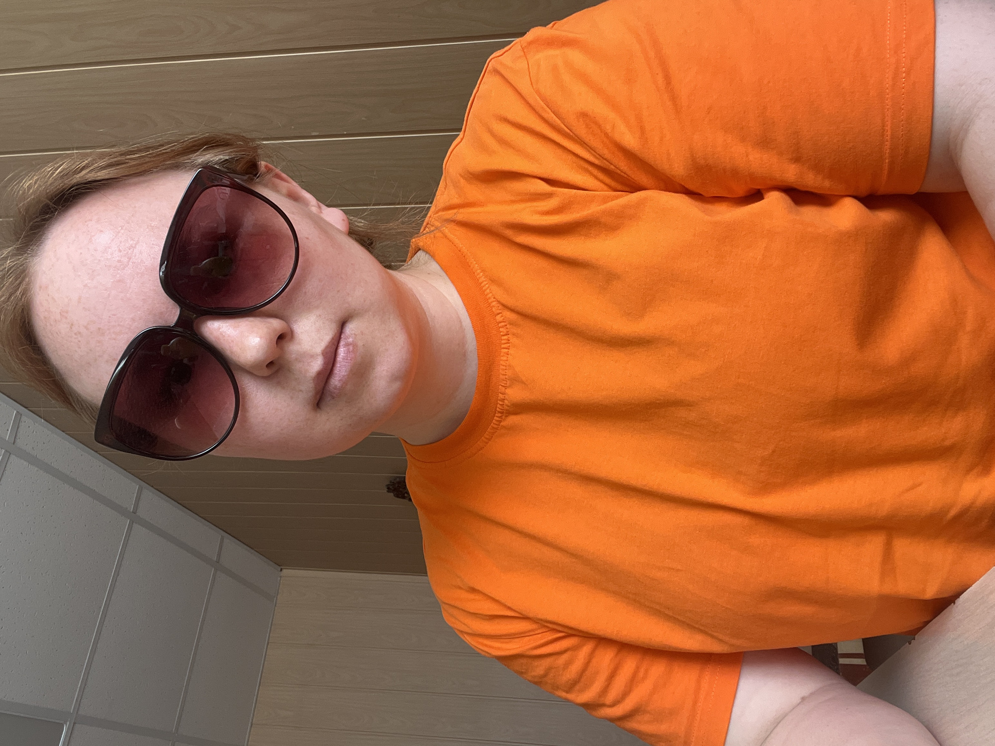A woman wearing an orange t-shirt and sunglasses is indoors. The background consists of a wall with light-colored horizontal paneling and another wall with large, light-colored tiles. The photo appears to have been taken from a low angle. There is no evidence to suggest the photo was taken in Chaikovsky, Russia.