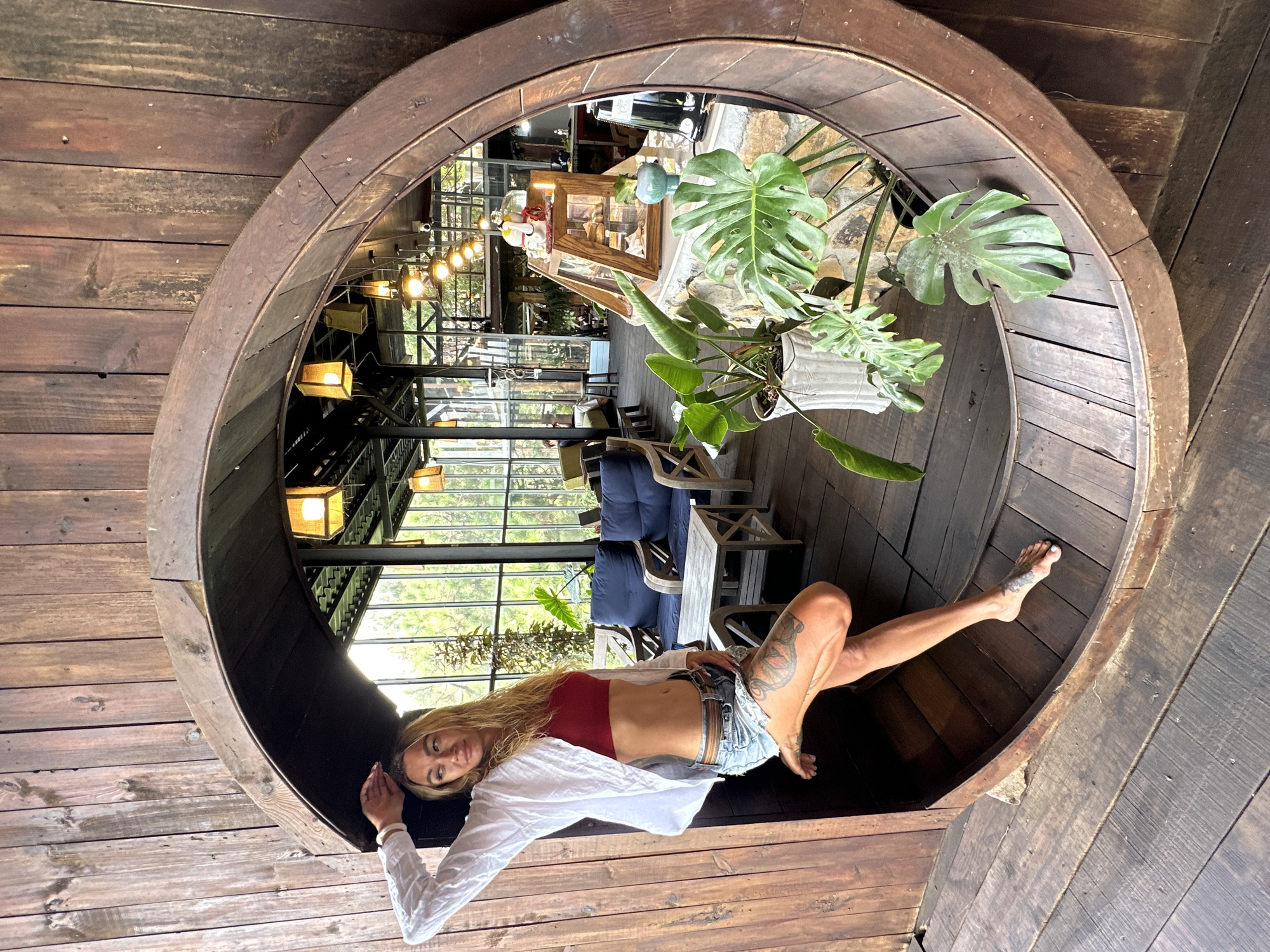 A woman reclines within a large, circular wooden structure, her body oriented horizontally in the frame. She wears a red bandeau top, denim shorts, and an unbuttoned white shirt. Tattoos are visible on her left leg. Her gaze is directed towards the camera, and she has a relaxed, somewhat serene expression.
The wooden structure appears to be part of a rustic, indoor setting, possibly a cafe or restaurant. It has a curved, barrel-like shape, with visible wood grain and nails. The interior reflected in the structure reveals a space with large windows, casting natural light. Inside, there are various objects, including plants, furniture such as tables and chairs, and overhead light fixtures. The overall impression is of a cozy, bohemian-style ambiance. The windows suggest it's daytime, and the lighting seems warm and inviting, hinting at a pleasant atmosphere. The setting is located in Da Lat, Vietnam, a city known for its cooler climate and natural beauty, which could explain the indoor-outdoor feel of the space.