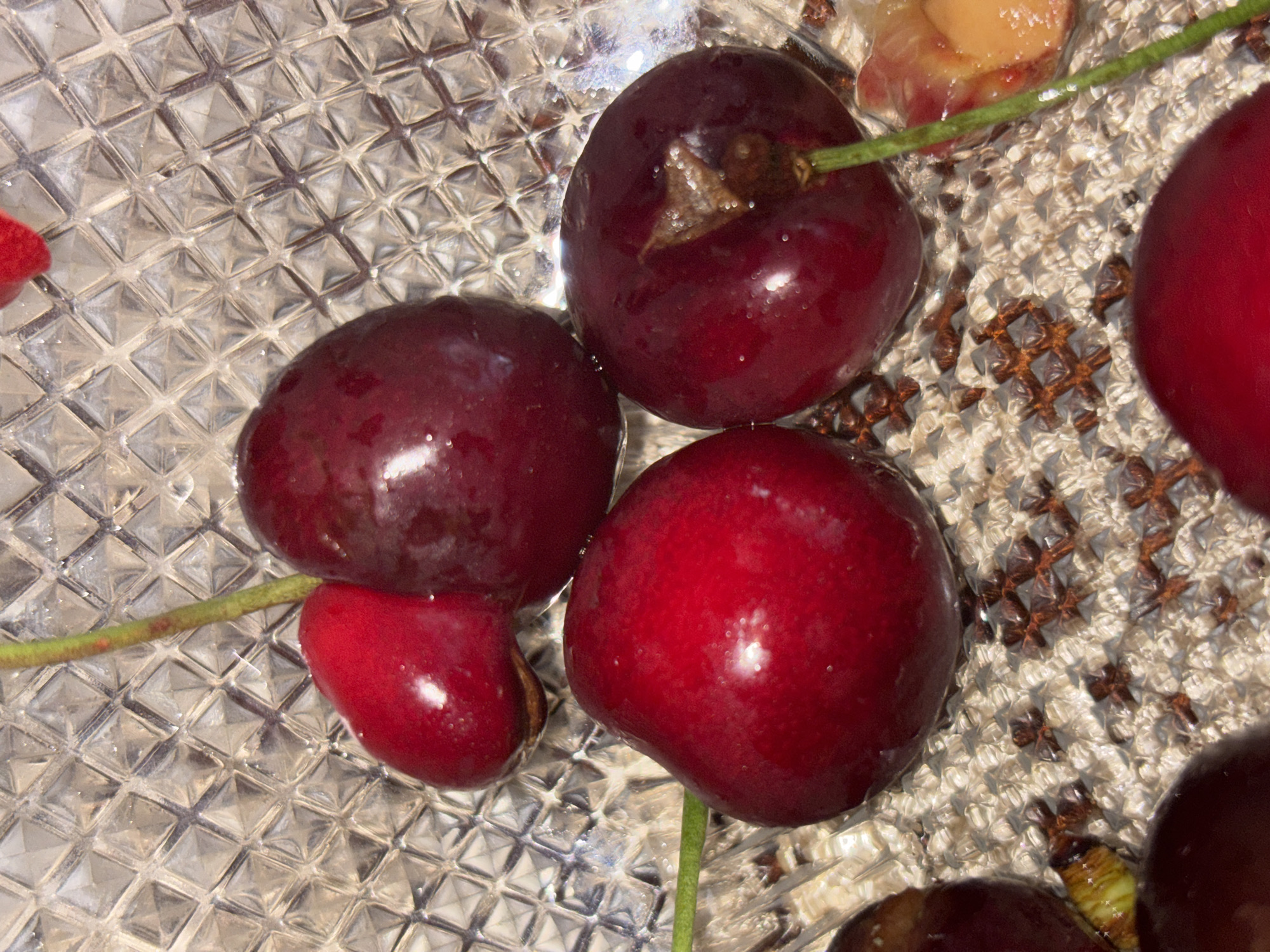 The image displays a close-up of several dark red cherries resting in a clear, textured glass dish. The dish features a distinctive grid pattern of raised squares, which creates numerous light reflections and refractions. Some cherries are still attached to their slender green stems, and many appear to be wet or glossy. One cherry is partially open or damaged, revealing its lighter, peach-toned flesh. In the background, a section of woven or embroidered fabric with a brown cross-stitch pattern is visible, likely serving as a placemat or tablecloth. The scene is static, set indoors under what appears to be natural or artificial lighting. No people, activity, specific time of day, weather, or visible text are present.
