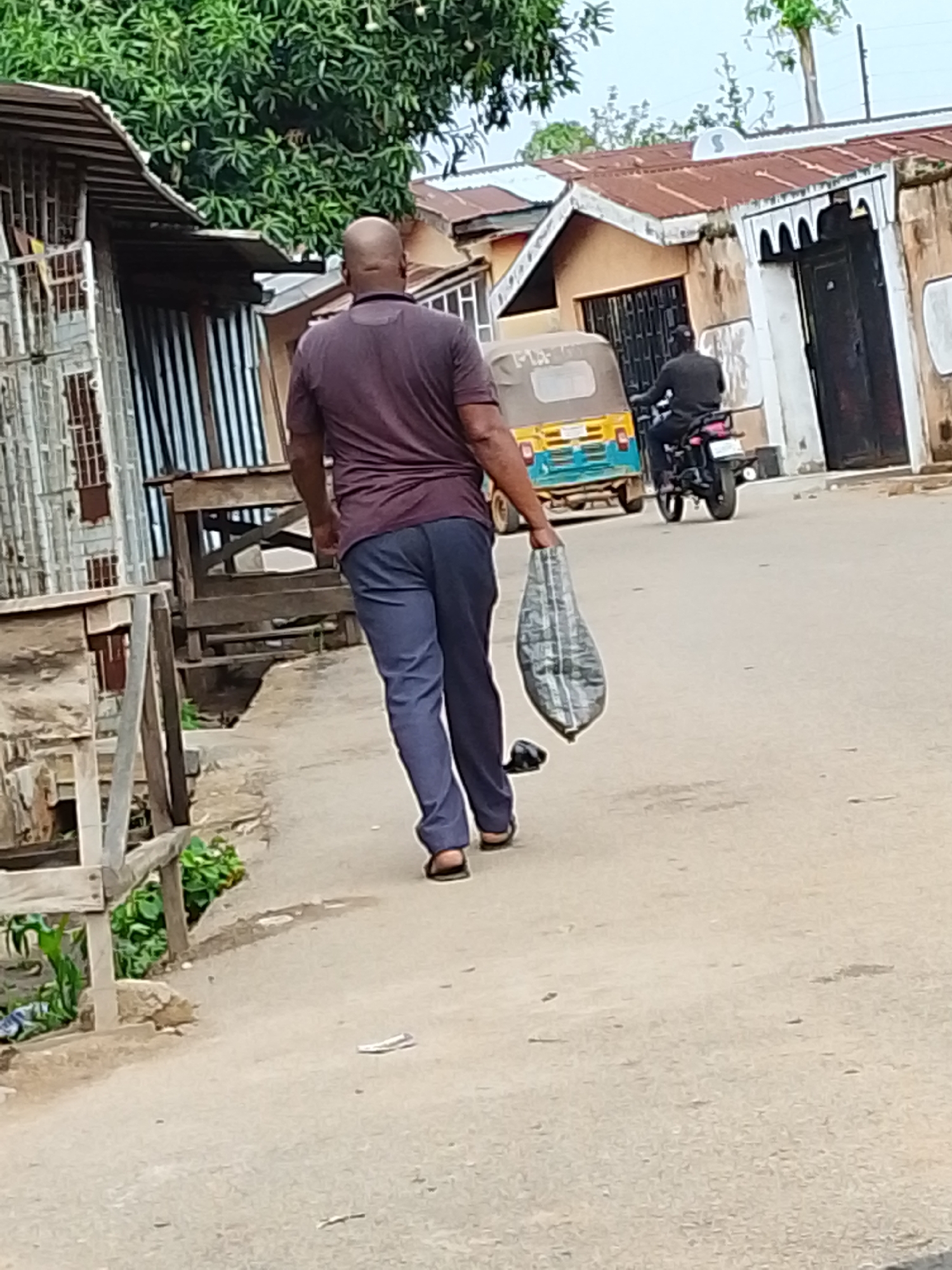 A bald, dark-skinned man in a maroon polo shirt and blue pants is walking away from the viewer on a paved road in what appears to be a city environment. He is carrying a grey patterned bag in his right hand. To his left is a wooden structure with green plants growing nearby. In the background, a yellow and blue three-wheeled vehicle and a motorcycle with a rider are on the road. Houses with rusty roofs and a black door are visible in the background. The location is the city of Unknown, Unknown.